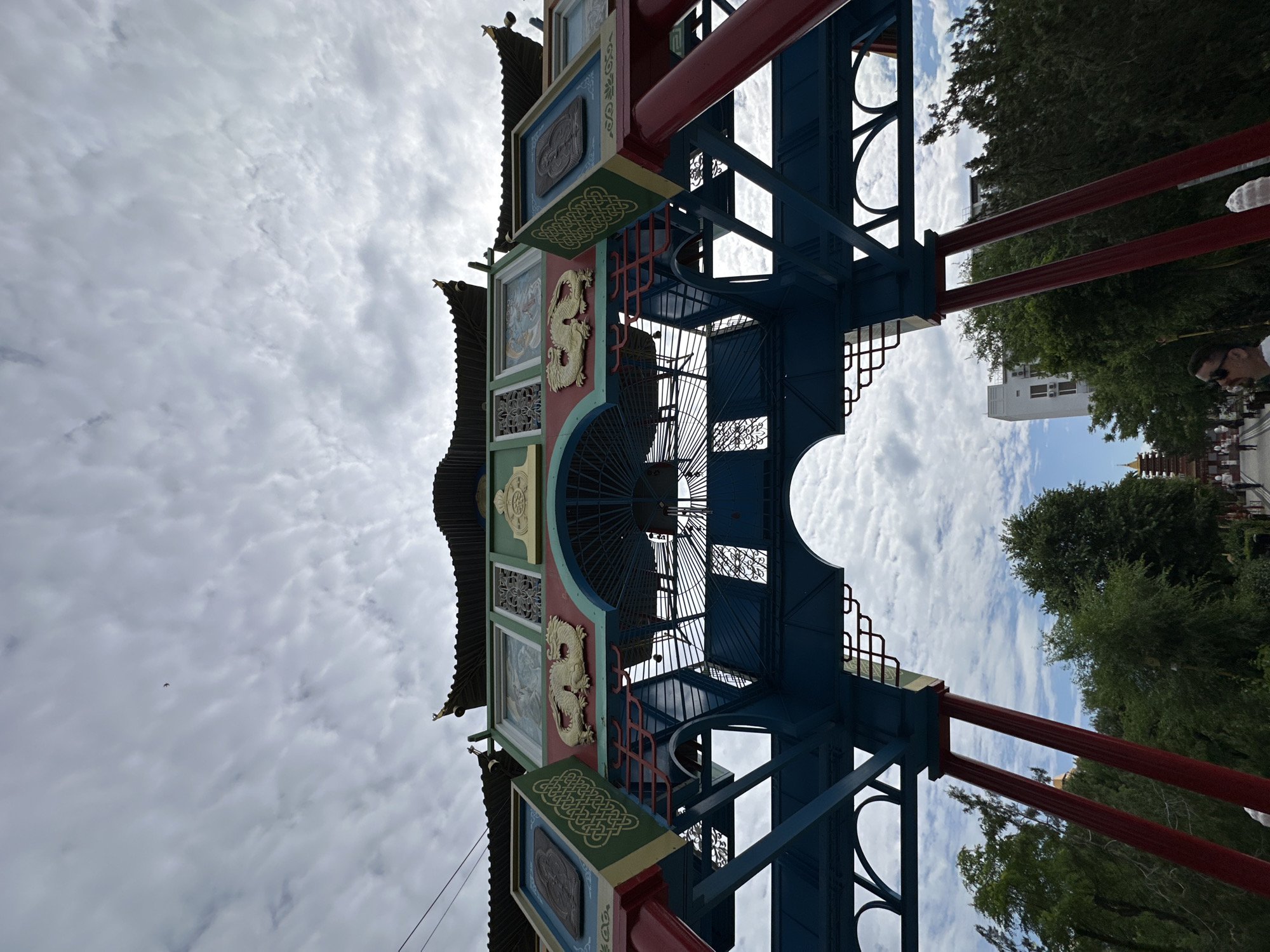 This is a low-angle, wide shot of a large, ornate Asian-style archway in Elista, Russia. The archway is painted in vibrant colors, with red supporting columns and blue structural elements. The top of the archway features traditional Chinese architectural details, including curved roofs, dragon motifs, and decorative panels. The sky above is overcast with scattered clouds, and a single bird is visible in the distance.
Through the archway, a street scene unfolds with lush green trees lining the path. In the background, several buildings are visible, including a modern white building and what appears to be a pagoda or temple-like structure with a golden roof. People are seen walking along the street, some appearing to be tourists. In the foreground, a man wearing sunglasses and a white shirt is looking towards the right side of the frame. The overall impression is of a cultural landmark situated within a city environment.