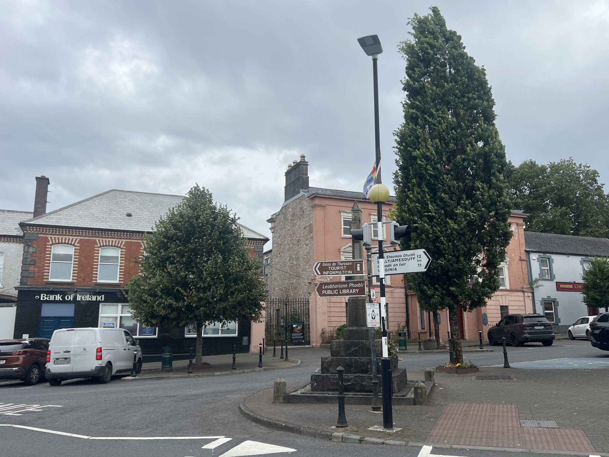 The image shows a street scene in an urban environment, possibly Ireland given the "Bank of Ireland" signage. The weather appears overcast. In the foreground, there is a road intersection with white lane markings. A large monument made of stone blocks is situated in the center of the intersection. A street lamp with a light fixture and a yellow globe is positioned next to the monument. A flag is attached to the street lamp.
Several buildings line the street. On the left, there is a dark-colored building with "Bank of Ireland" in white lettering and blue accents. Next to it, a lighter-colored building with brick detailing and large windows. A white panel van is parked in front of the bank. To the right of the monument, a salmon-pink building is visible, with signage indicating directions to "TOURIST INFORMATION" and "PUBLIC LIBRARY." Further to the right, a grey building with blue trim is visible, and a sign that says "TAKE OUT CHI".
Trees are present on both sides of the street, with a particularly tall, slender evergreen tree dominating the right side of the frame. Several cars are parked or driving in the scene, including a dark SUV and a white van. The ground is paved with asphalt and cobblestones. There are also several black bollards placed along the sidewalks and around the monument. The overall impression is a town square or intersection in a moderately busy commercial area.