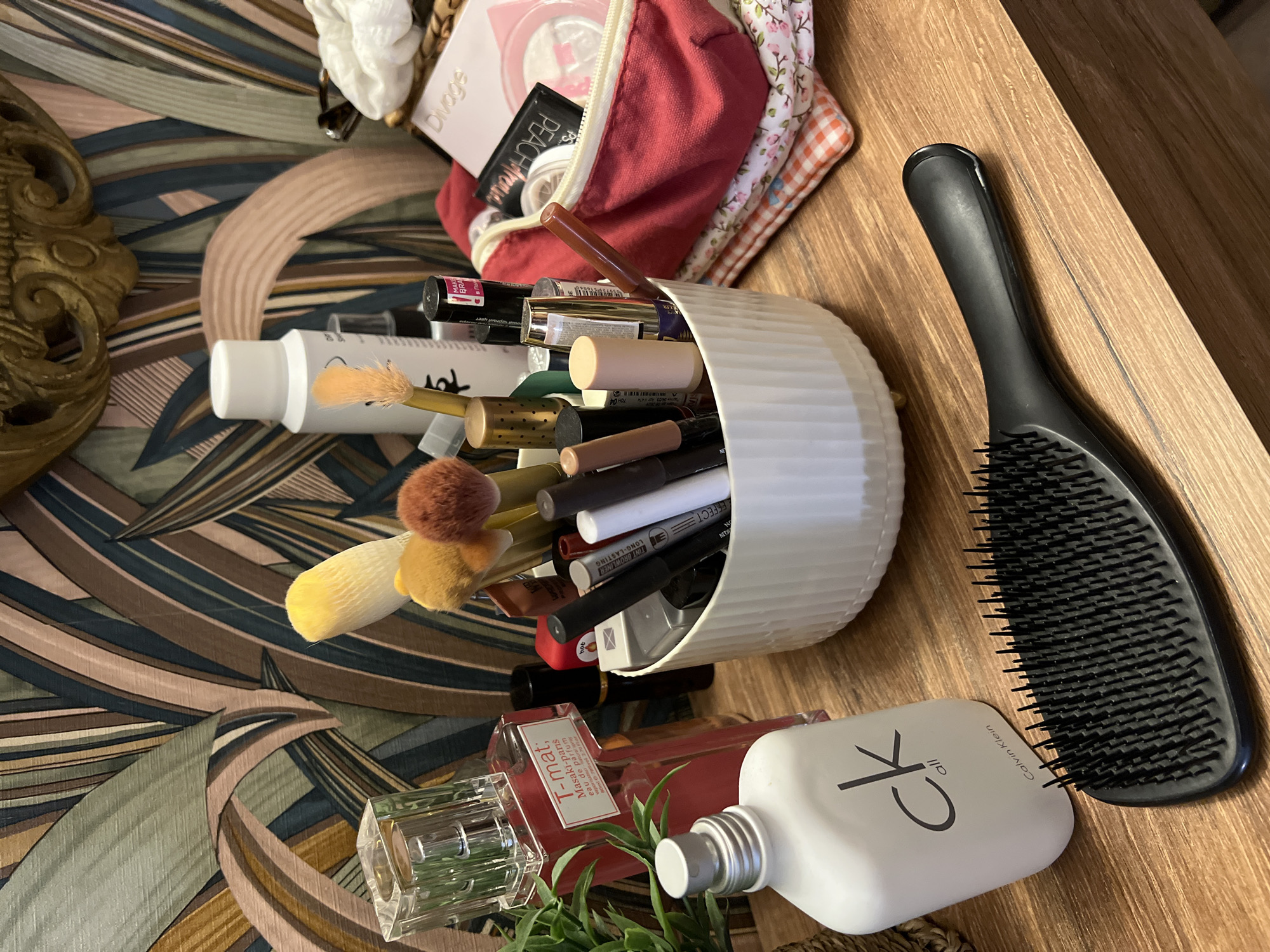 An overhead shot shows a wooden dresser top adorned with various beauty products and accessories. In the foreground, a black detangling hairbrush lies horizontally, its bristles pointing upwards. To the left of the brush, a white bottle with the "ck all" logo and "Calvin Klein" text is positioned upright. Behind these items, a clear glass perfume bottle with a pink liquid and a white label reading "T-mat Masaki Matsushima eau de parfum" stands to the left. Adjacent to the perfume bottle is a white cylindrical holder filled with makeup brushes and various cosmetics, including lipsticks, mascaras, and eyeliners. To the right of the holder, a red makeup bag is partially open, revealing makeup products and a white scrunchie peeking out from a woven basket behind it. The background features a patterned wallpaper with large, abstract leaf motifs in muted tones of green, brown, beige, and pink. A golden, ornate mirror frame is visible at the top center of the image. The overall scene suggests a personal vanity or dressing area.