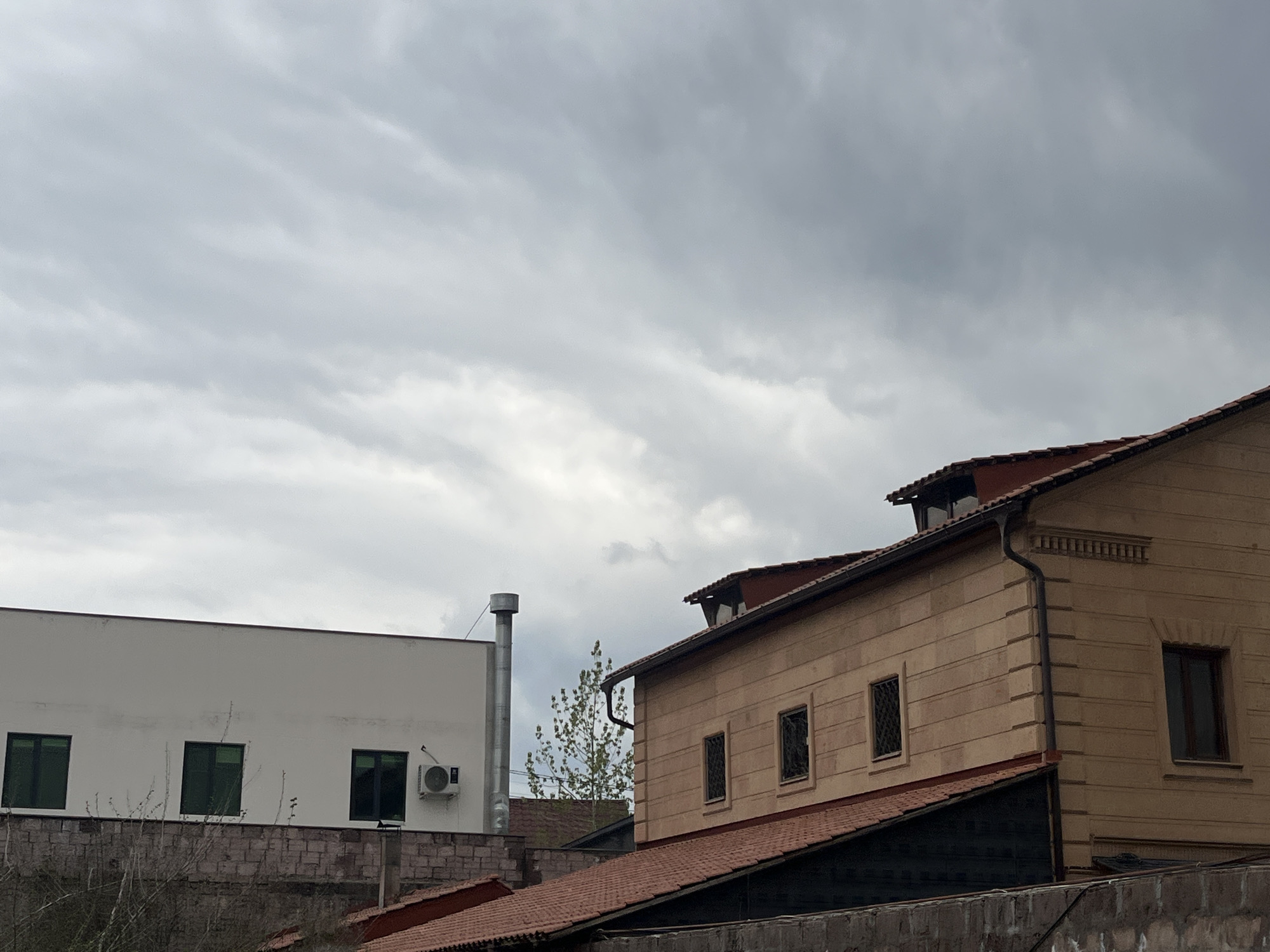 The photograph depicts two buildings under an overcast sky in Yerevan, Armenia. The building on the left is a modern, white, rectangular structure with three green-framed windows and an air conditioning unit. A metal chimney is visible next to the building. In front of it, is a low stone wall. The building on the right is a multi-story, beige-colored structure with dark windows and a red-tiled roof. The roof extends down to a lower level, which also has a dark-colored wall. In the middle of the two structures is a sparse tree. The overall scene is urban and slightly gloomy due to the weather.
