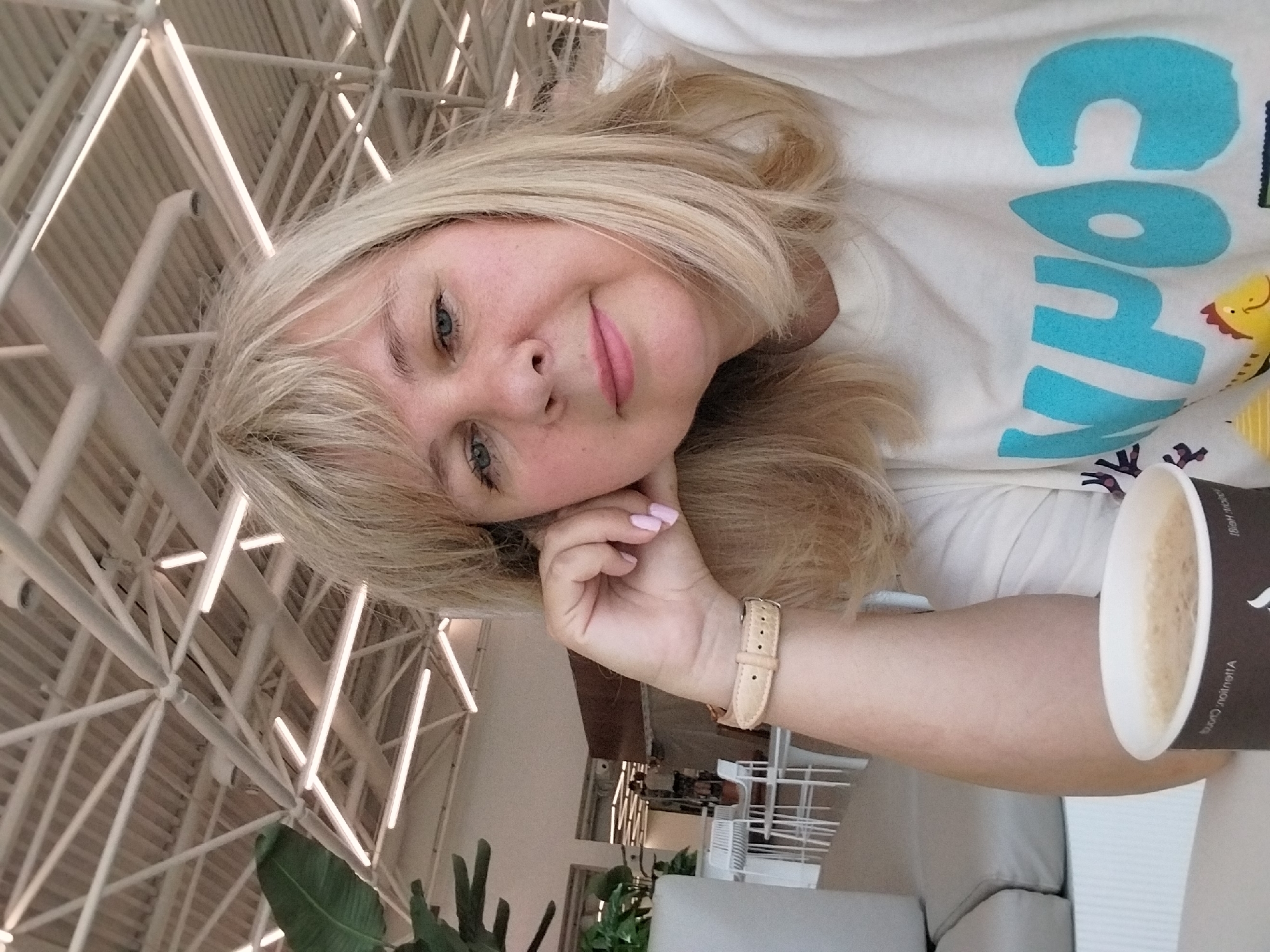 A blonde woman with blue eyes and light pink nail polish is taking a selfie in what appears to be a brightly lit cafe or restaurant. She is resting her left arm on a table and leaning her head on her hand, with a cup of coffee in front of her. She is wearing a white t-shirt with large blue lettering and a cartoon image. In the background, there is a modern ceiling with exposed beams and lighting fixtures, as well as some tables and chairs. There is also a large potted plant on the left side of the image. The woman's location is not specified in the image or context, therefore it is not possible to determine if she is in Orenburg, Russia.