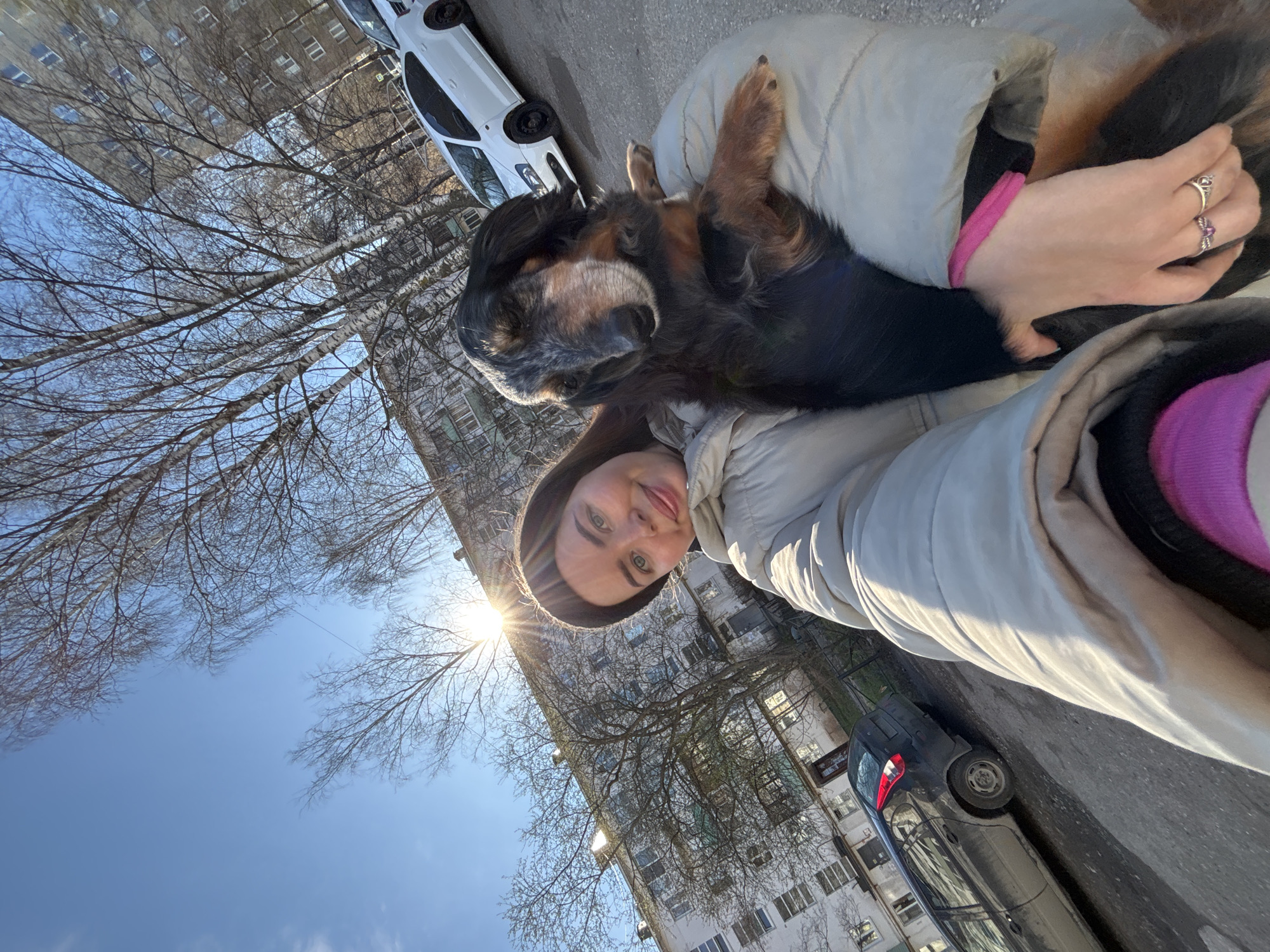 The image is a tilted selfie featuring a young woman and her dog outdoors in Izhevsk, Russia, on a bright, sunny day. The woman, with medium-length brown hair and a subtle smile, wears a light-colored puffer jacket, a dark beanie, and a bright pink sleeve or cuff is visible. Rings are visible on her right hand. She is holding a long-haired dog, which appears to be an older breed with a black and brown coat and significant graying around its muzzle. The dog looks calm and is resting against her, wearing a pink collar.
The background reveals a clear blue sky with a prominent sunburst on the left side, indicating bright sunshine. Bare deciduous trees with many intricate branches, typical of winter or early spring, frame the upper and middle sections of the scene. Behind the subjects and trees are tall, light-colored apartment buildings with numerous windows, suggesting a residential area. A paved street with parked cars, including a white car and a dark sedan, is visible towards the bottom and right of the frame. No discernible text is clearly visible on any objects in the scene. The overall mood is cheerful and relaxed.