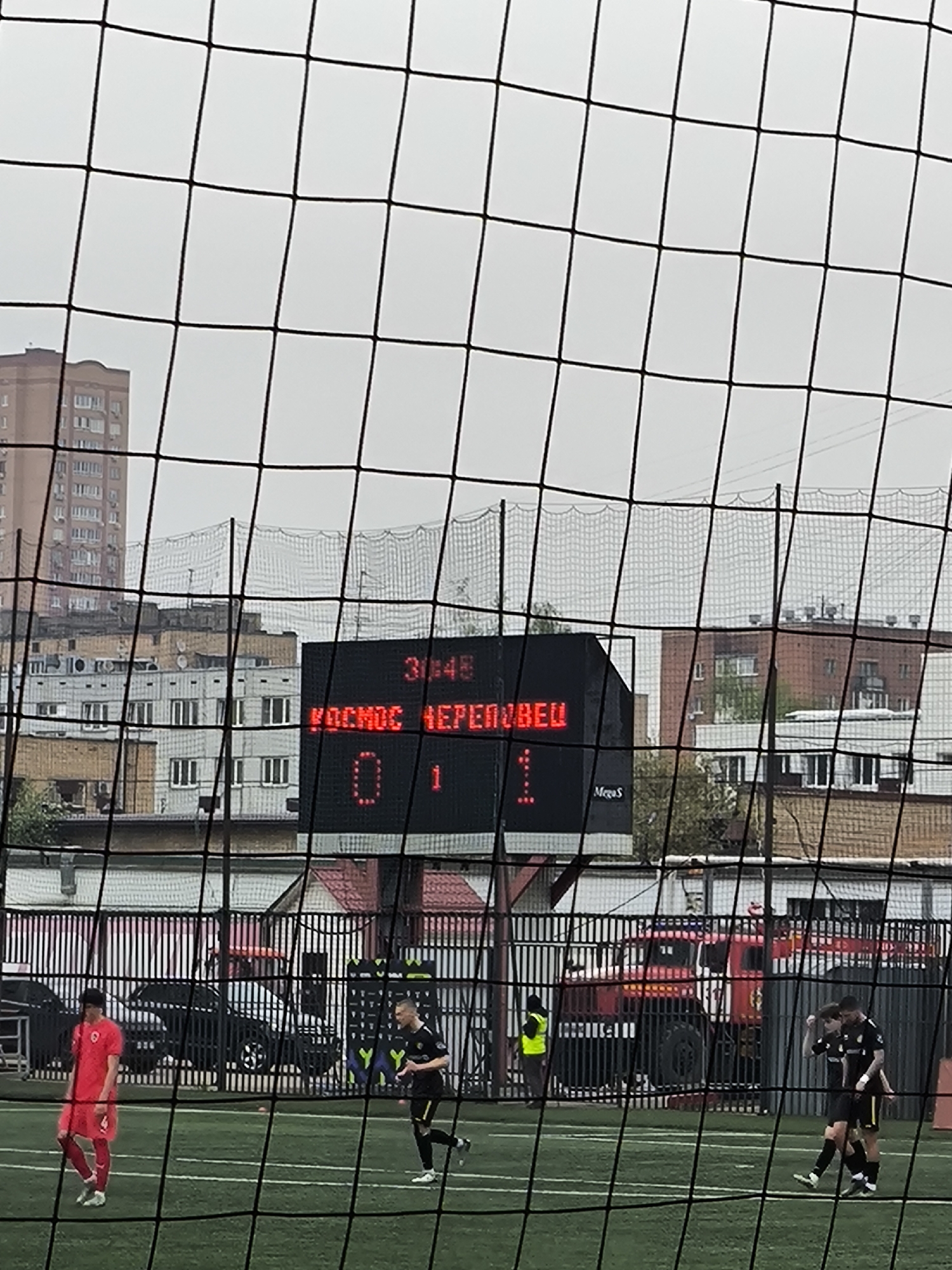 The image depicts a soccer field scene, likely in Khimki, Russia, as the digital scoreboard displays "KOSMOS CHEREVEZ" (translated as Cosmos Cherevez), the name of a soccer team in Khimki. The score is 0-1, with the game clock at 31:45. Several apartment buildings are in the background, typical of an urban environment. A black chain-link fence separates the field from the background.
On the field, several soccer players are visible. A player wearing a red jersey is on the left side of the frame, and two players in black and yellow jerseys are on the right. Another player in black and yellow is mid-stride towards the center.
Behind the fence, various vehicles, including black cars and a red fire truck, are present. A person in a bright yellow safety vest stands near the fire truck. The sky is overcast. A soccer net obscures some of the foreground.