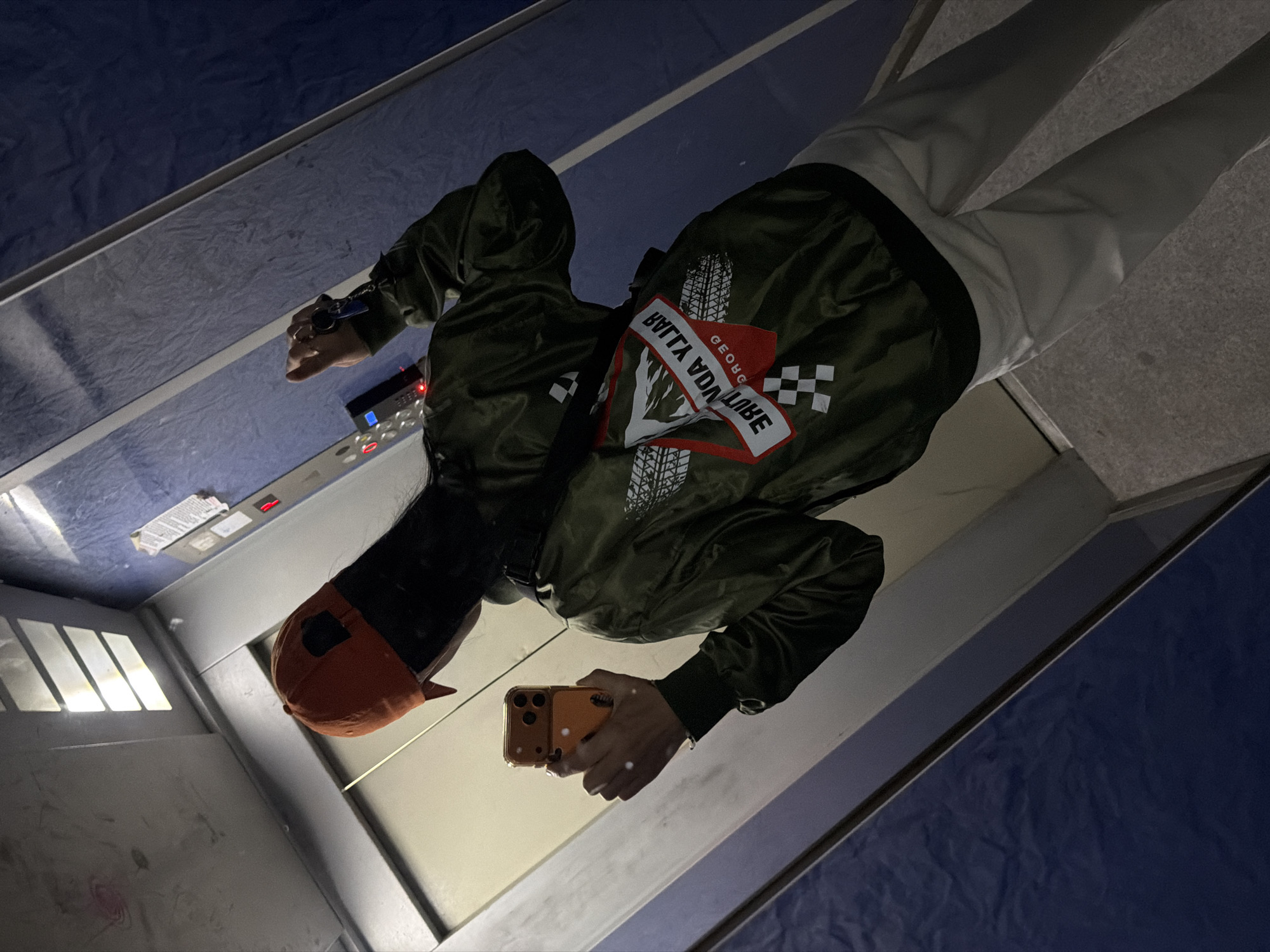 A person is taking a selfie in what appears to be an elevator. The person is wearing white pants, a green jacket with an orange cap. The reflection shows the elevator control panel, and a partially visible ventilation grid. The interior of the elevator and the blue walls of the building suggest the location is in Tbilisi, Georgia. The lighting suggests it is taken at night or in a dimly lit area.