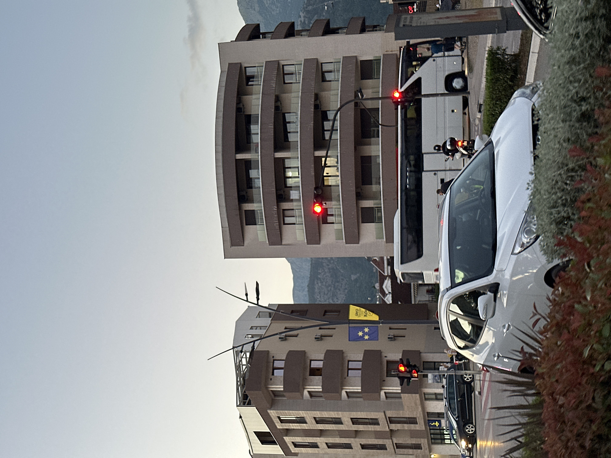 The image depicts a street scene in Budva, Montenegro. Two modern apartment buildings with curved balconies are visible. A large white bus is stopped at a traffic light, and a motorcycle with a rider is alongside it. In the foreground, a white SUV is partially obscured by greenery. Traffic lights are red, indicating a stop. Two flags are hanging from a pole on the left building; one is blue with yellow stars, and the other is yellow with text. Mountains can be seen in the background under a hazy sky.