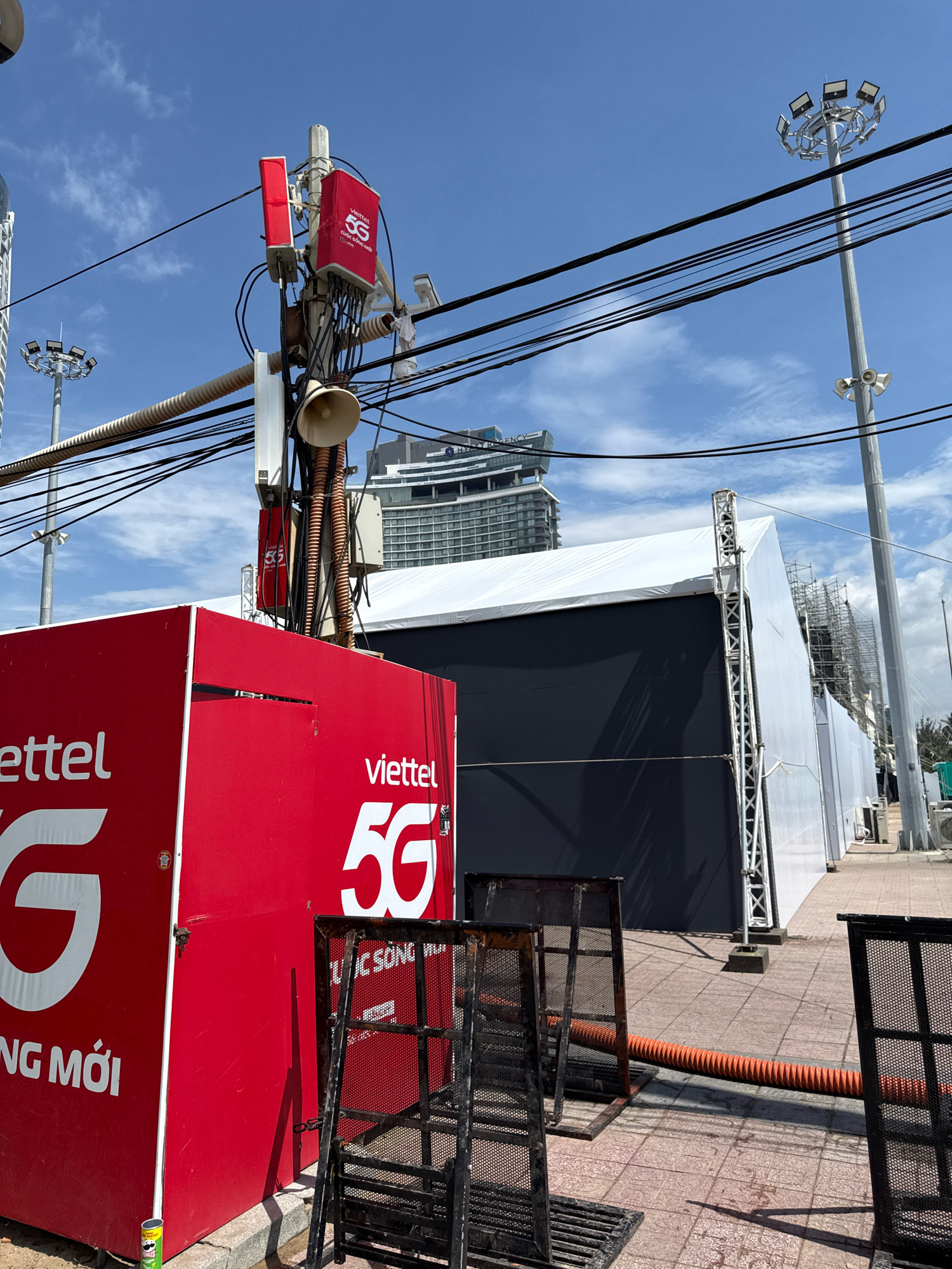 This image depicts a street scene in Nha Trang, Vietnam, likely in a public area or event space. In the foreground, a bright red, rectangular structure, possibly a generator or storage unit, is prominently displayed with the "Viettel 5G" logo in white text. The structure also has a large white "G" symbol. To the right of this structure, several black metal barriers are set up, some connected by an orange corrugated pipe.
Behind the red structure, a large white tent with dark panels forms a temporary building or stage. A tall telecommunications tower with red "Viettel 5G" antennas and various cables is visible against a bright blue sky with scattered clouds. Further in the background, a tall building with a modern architectural design stands, along with a large street lamp with multiple spotlights. The ground appears to be paved with tiles, and the overall impression is that of an outdoor event or construction site. There are no people clearly visible in the image, but the presence of event infrastructure suggests a public gathering or temporary setup.
