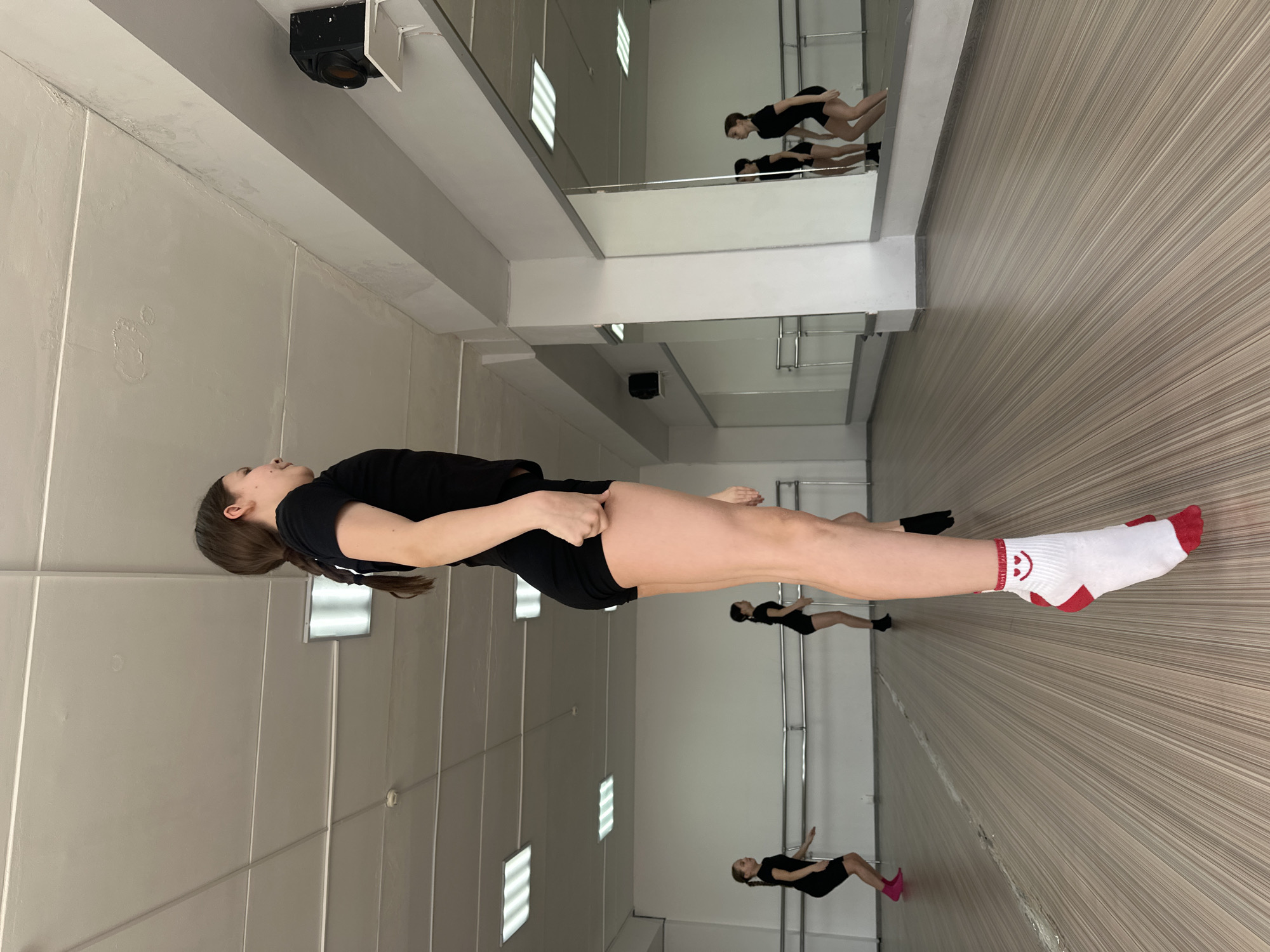 A young woman in black athletic wear and one white sock with a red toe and heel stands in a dance studio. The studio has wood-look flooring and gray paneled walls with several windows. A mirror runs along part of the wall, reflecting the woman and the studio. There is no indication that this image was taken in Votkinsk, Russia.