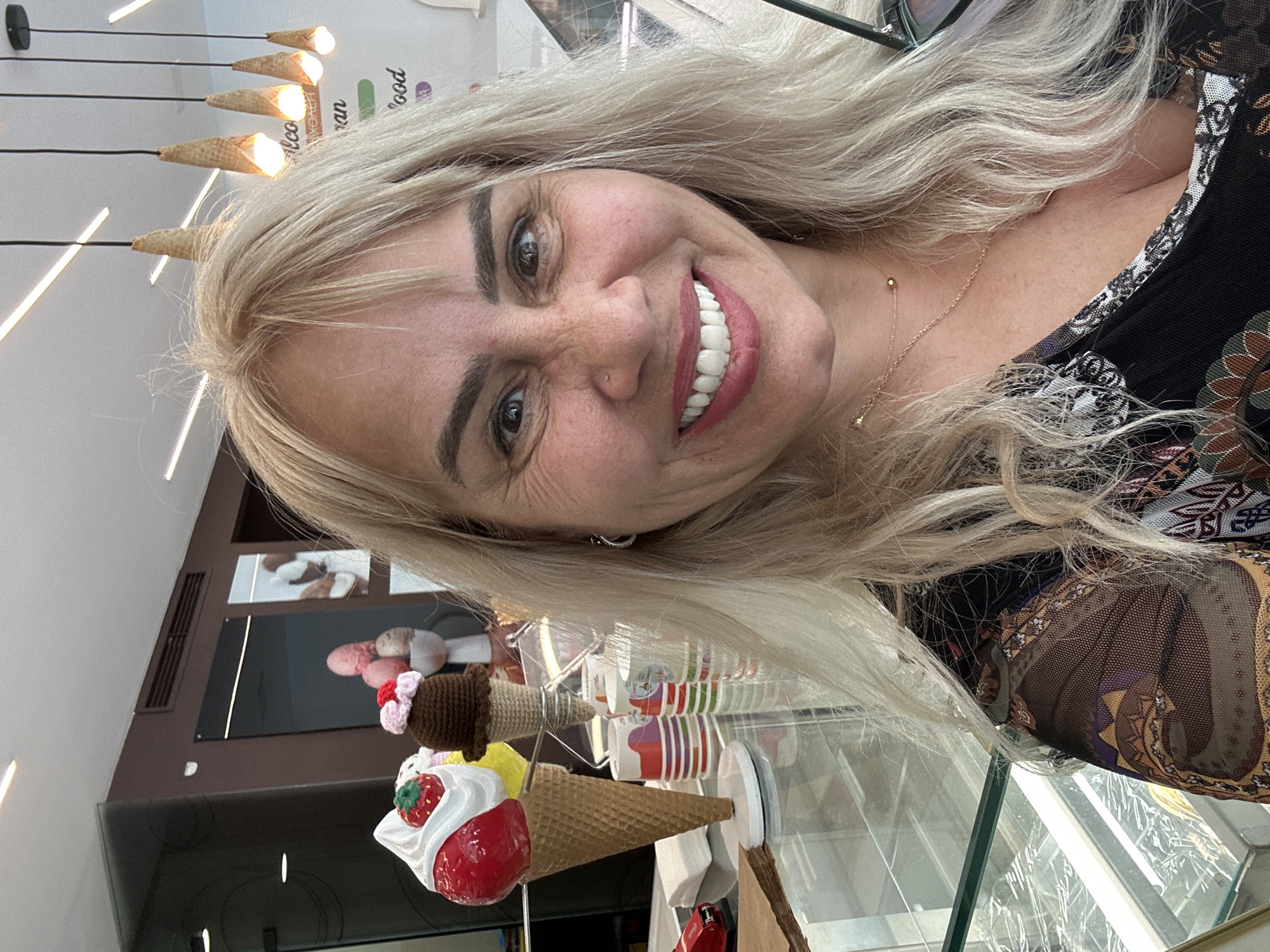 A cheerful woman is captured in a close-up, selfie-style photograph, smiling broadly into the camera. She has long, wavy blonde hair, dark eyes, and is wearing a patterned top with intricate designs. A delicate gold necklace with small beads and a nose piercing are visible. Sunglasses rest on top of her head.
The setting appears to be an ice cream parlor or dessert shop, likely located in Sofia, Bulgaria. The background features several distinct ice cream-themed decorations, including five hanging ceiling lights shaped like inverted ice cream cones, illuminating a light-colored wall. A large, realistic prop ice cream cone, topped with a red strawberry, white whipped cream, and yellow ice cream, is prominently displayed in front of a glass counter. Smaller, crocheted ice cream cones and stacks of colorful paper cups are also visible in the display area.
Partially visible text on the wall behind her includes fragments like "Leo," "ran," and "ood," suggesting part of a brand or shop name. The bright indoor lighting indicates the photo was taken during the day, and the woman's clear, wide smile conveys a joyful and friendly demeanor.
