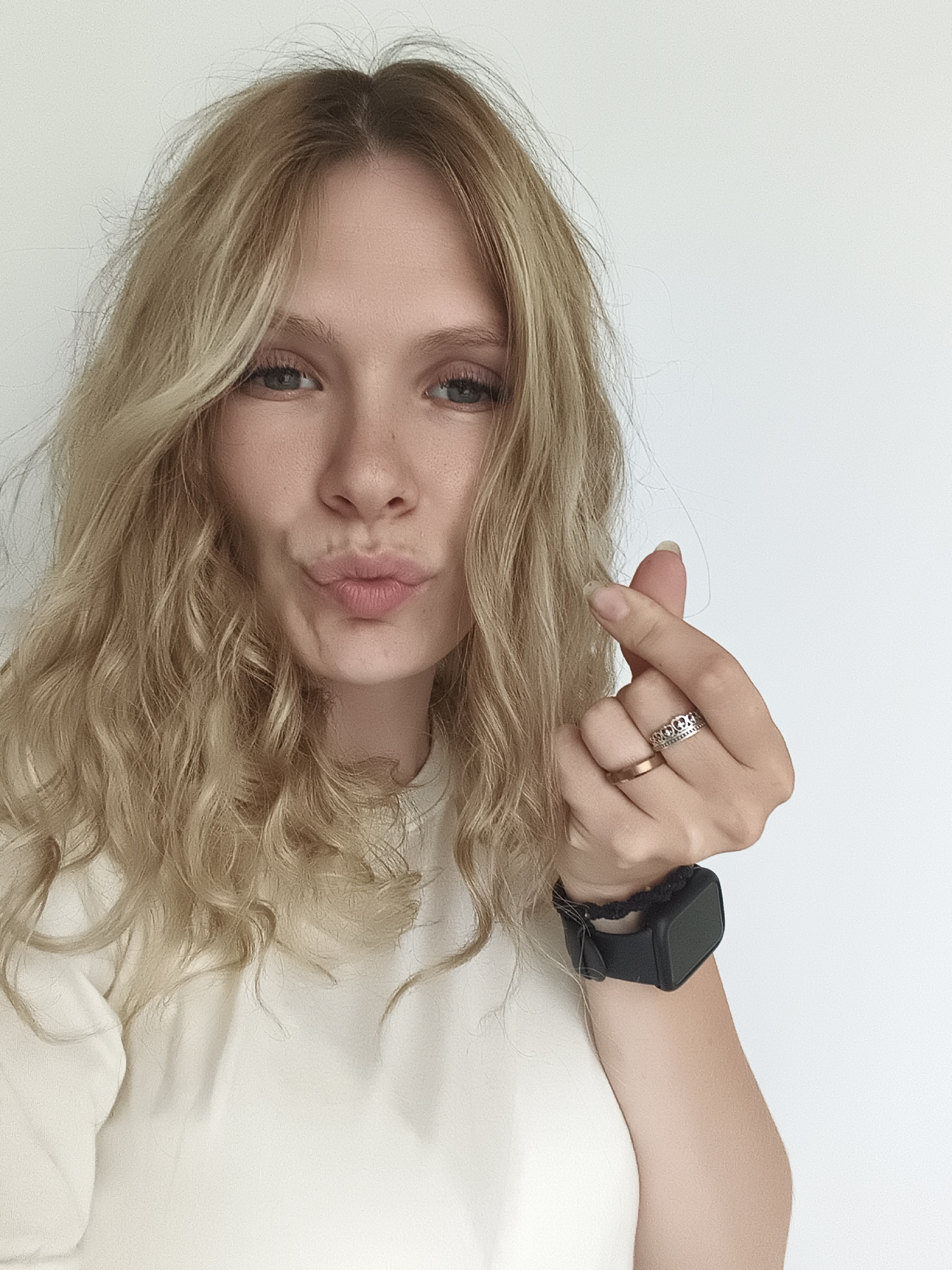 A young woman with blonde, wavy hair is looking towards the camera and making a heart shape with her thumb and index finger. She is wearing a light-colored long-sleeved shirt and a black smartwatch. Her lips are pursed in a kiss. The background is a plain white wall. There is no identifiable information about the location in Izhevsk, Russia.