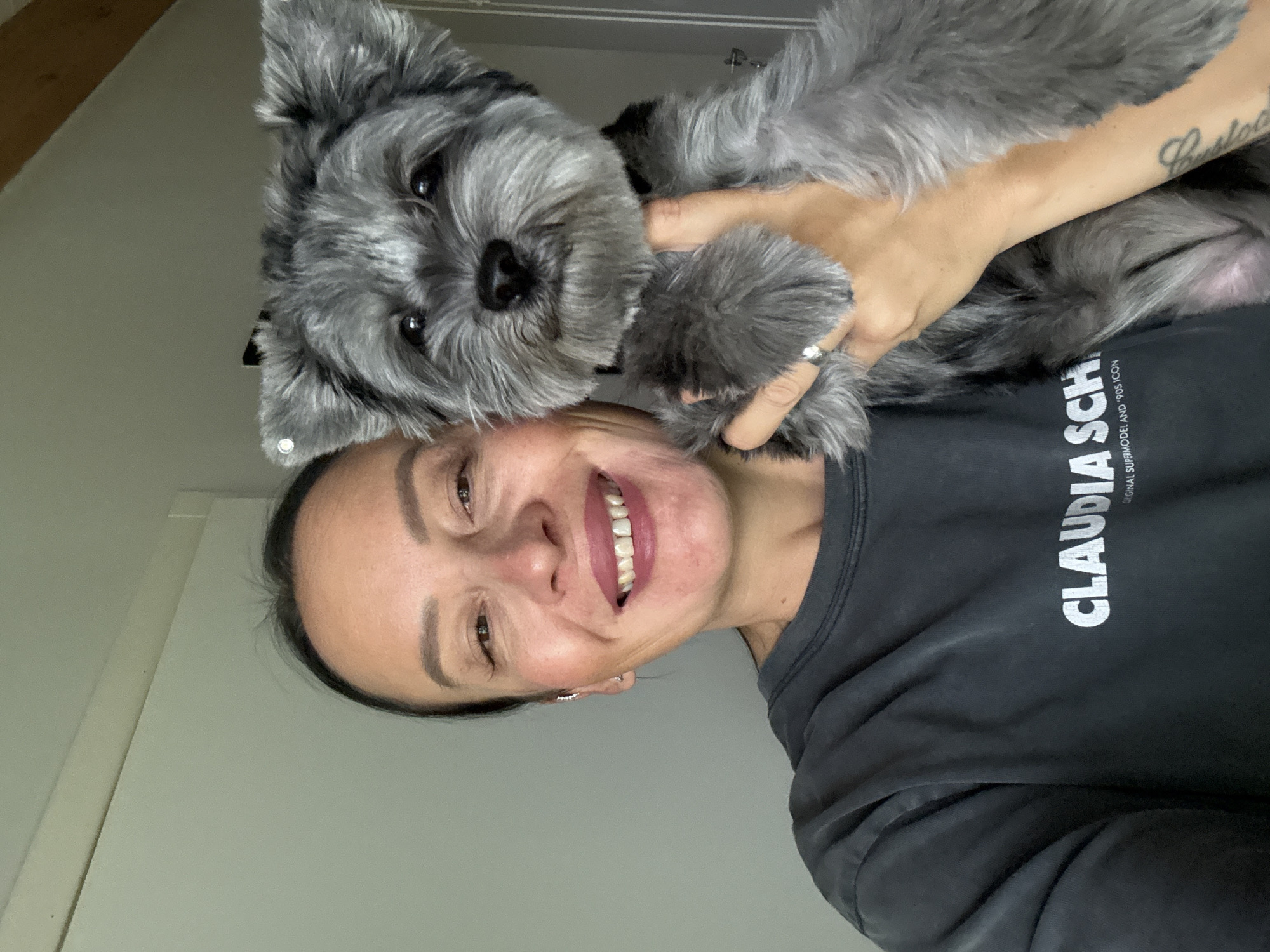 A medium shot shows a woman holding a small, gray, shaggy dog. The woman is smiling and looking at the camera. She has dark hair pulled back and is wearing a dark gray t-shirt that says "CLAUDIA SCH" in white letters. The dog has dark eyes, a black nose, and pointed ears. The background is a plain, light-colored wall with some molding at the top. There is also a hint of a wooden beam in the upper right corner. The lighting suggests an indoor environment, possibly a home. The location of Chaikovsky, Russia, is not discernable from the image.