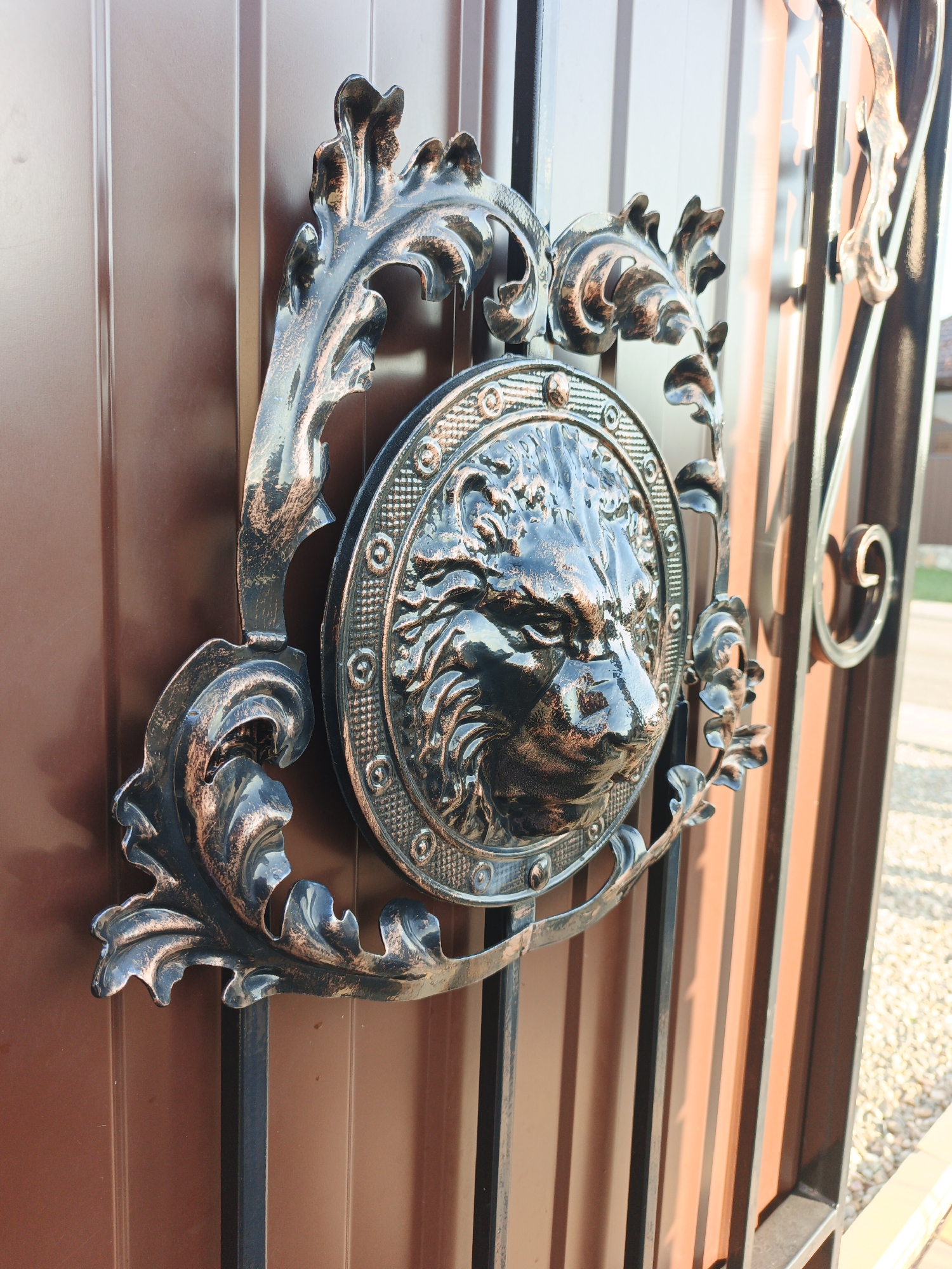 A close-up, eye-level shot captures a section of a brown metal gate featuring an ornate, wrought-iron decorative element. This element is centered around a circular medallion depicting a detailed, fierce wolf's head with flowing mane and mane. The medallion is surrounded by a decorative frame that incorporates stylized leaf motifs and swirling patterns. The metalwork is primarily dark with a coppery patina highlighting the raised textures and edges. The gate itself is composed of vertical brown panels. To the right of the central decoration, vertical black metal bars of the gate are visible, with additional scrolling metalwork. Sunlight casts shadows across the gate and the metallic elements, suggesting an outdoor setting. The background is blurred, showing hints of gravel and possibly a building or fence, indicative of a residential or public area. The location is identified as Repnoe, Russia.