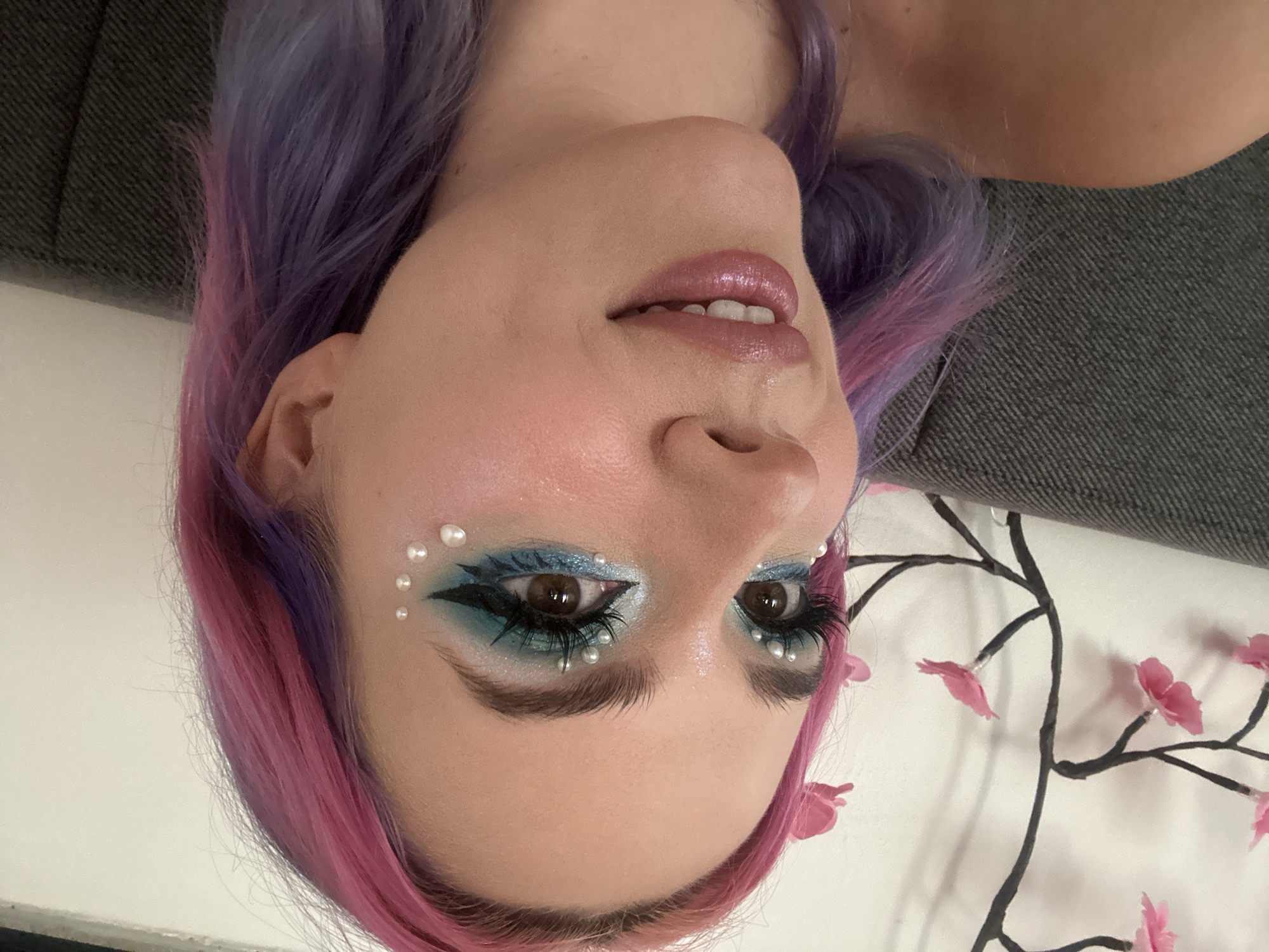 A close-up, upside-down shot depicts a person with pink and purple hair. The individual is adorned with distinctive makeup, featuring blue eyeshadow accented with small pearl-like beads around the eyes and temple. The lips are glossy, and the skin has a dewy finish. The person is lying on what appears to be a bed with a floral pattern. A gray couch is visible in the background. There is no direct evidence to suggest this image was taken in the City of Zagreb, Croatia.