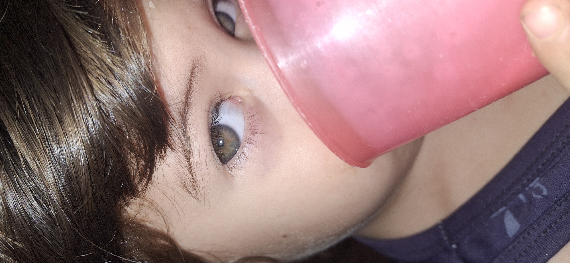 A close-up shot shows a child drinking from a pink cup. The child has brown hair and brown eyes. The child is wearing a dark blue shirt. The child's face is fair-skinned. The background is out of focus. It is not possible to determine the location of the image based on the information provided.