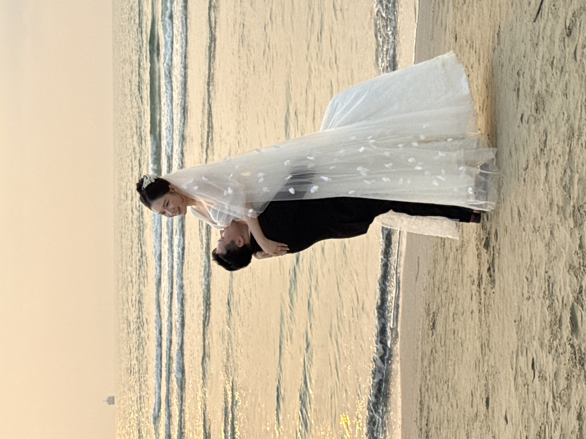 A couple is in the middle of a wedding photoshoot on the beach in Đà Nẵng, Vietnam. The bride is wearing a white wedding dress and veil with floral embellishments, and the groom is wearing a black suit. The groom is holding the bride in a dip pose. They are positioned near the shoreline. The water is lapping at the sand, and the sky is a muted color, likely indicating either dawn or dusk. The background shows the ocean meeting the horizon, and a building can be seen far in the distance. The sand is grainy and light colored, typical of a beach environment.