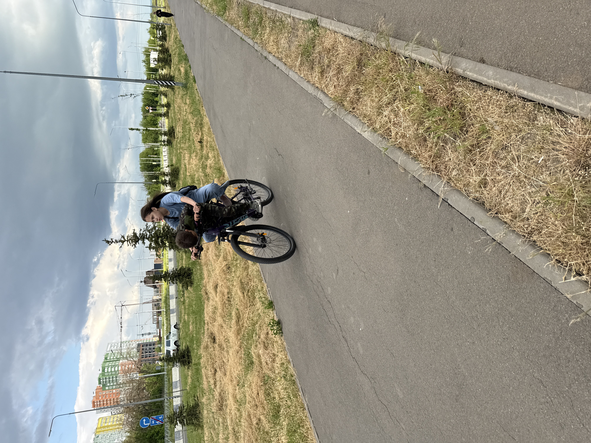 A medium shot, taken at eye level, presents a scene of two individuals on a bicycle path in Rostov-on-Don, Russia.
In the foreground, a woman with dark hair is positioned on the back of a bicycle. She is leaning forward and holding onto the shoulders of a younger person, presumably a child, who is riding the bicycle. Both figures are facing forward along the path.
The bicycle path itself stretches into the distance, bordered by a strip of dry grass. Beyond this, the urban landscape of Rostov-on-Don is visible. There are buildings of varying heights and colors, suggesting a modern city environment. Streetlights punctuate the scene, and the sky above is a mix of blue and overcast clouds.
The image composition emphasizes the act of riding a bicycle on the path, set against the backdrop of a city. The presence of a younger person being assisted by the woman suggests a learning or recreational activity.