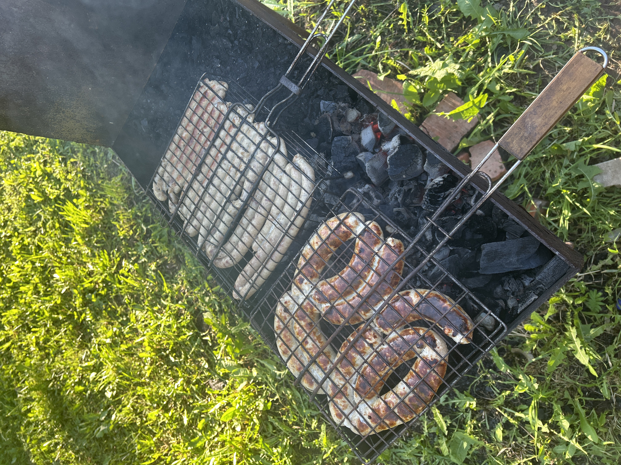 The image shows a barbecue grill with various sausages cooking over charcoal. Some sausages are pale, indicating they are still raw or only partially cooked. Others have a darker, grilled appearance. The grill is set on grass. The location cannot be confirmed as Фоки, Russia based on visual evidence alone.