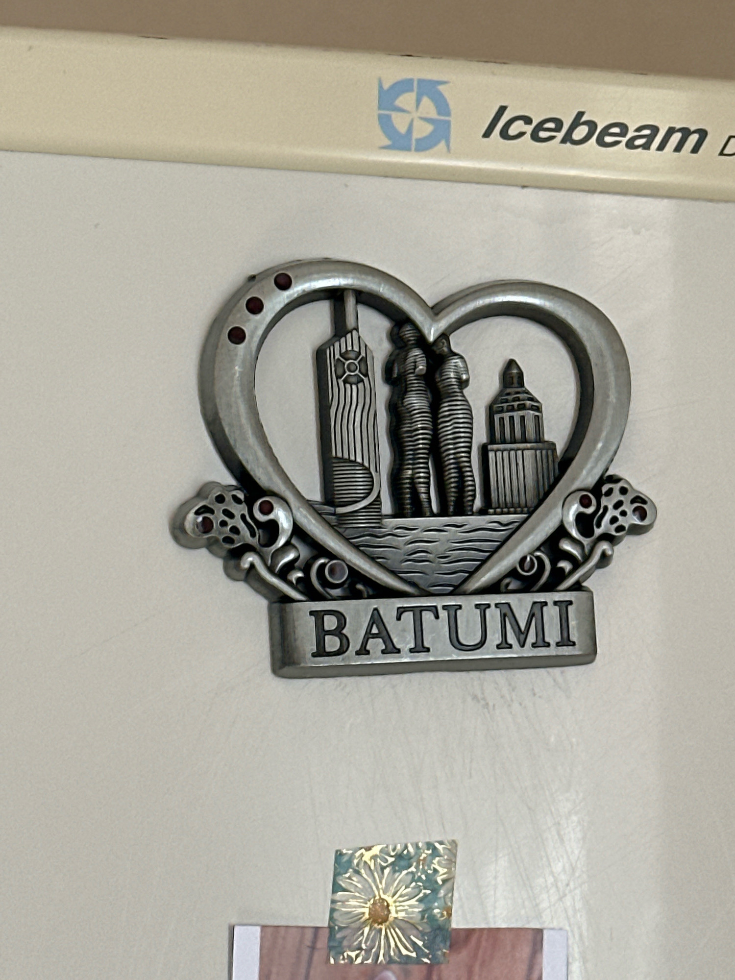 The image shows a metallic heart-shaped refrigerator magnet with the word "BATUMI" written on a rectangular plaque attached to the bottom. The magnet features a depiction of several buildings and statues, possibly representing landmarks in Batumi, Georgia. The magnet is attached to a light-colored surface, likely a refrigerator door, and has a faint imprint of the "Icebeam D" logo in the upper right corner. Below the magnet, a small square sticker with a floral design is partially visible. The overall impression is of a souvenir item displayed on an appliance. The image does not depict any people or suggest any specific location within Gyumri, Armenia, as Batumi is a city in Georgia.
