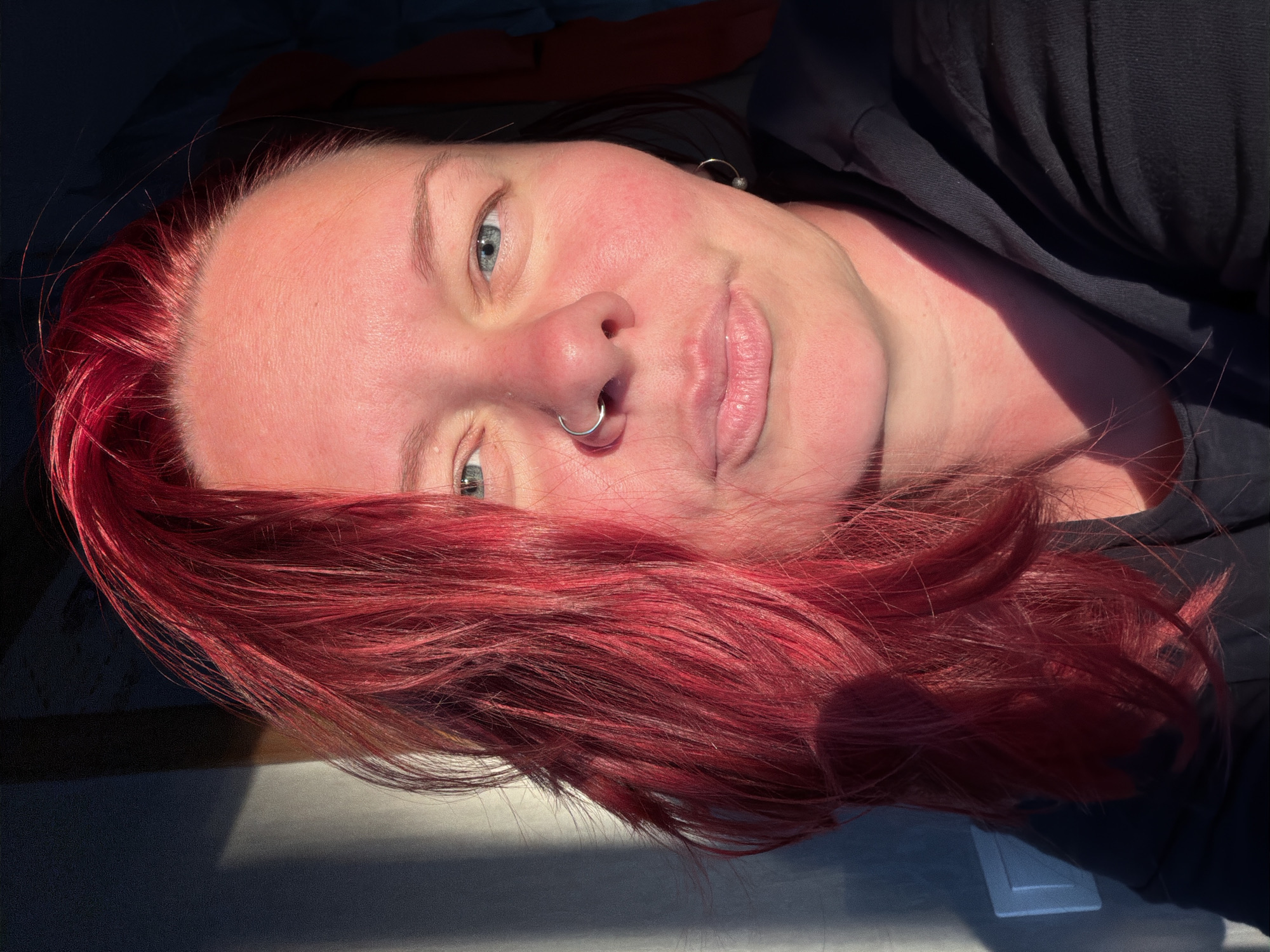 A woman with bright red hair and blue eyes looks directly at the camera in a close-up shot. She is wearing a silver hoop nose ring and a black hoodie. The lighting suggests she is outdoors during the golden hour, with strong shadows cast on the wall behind her, creating a dappled effect. The background is dark and out of focus, with a hint of a wall with a light switch on the left. No specific location in Chaikovsky, Russia is discernible.