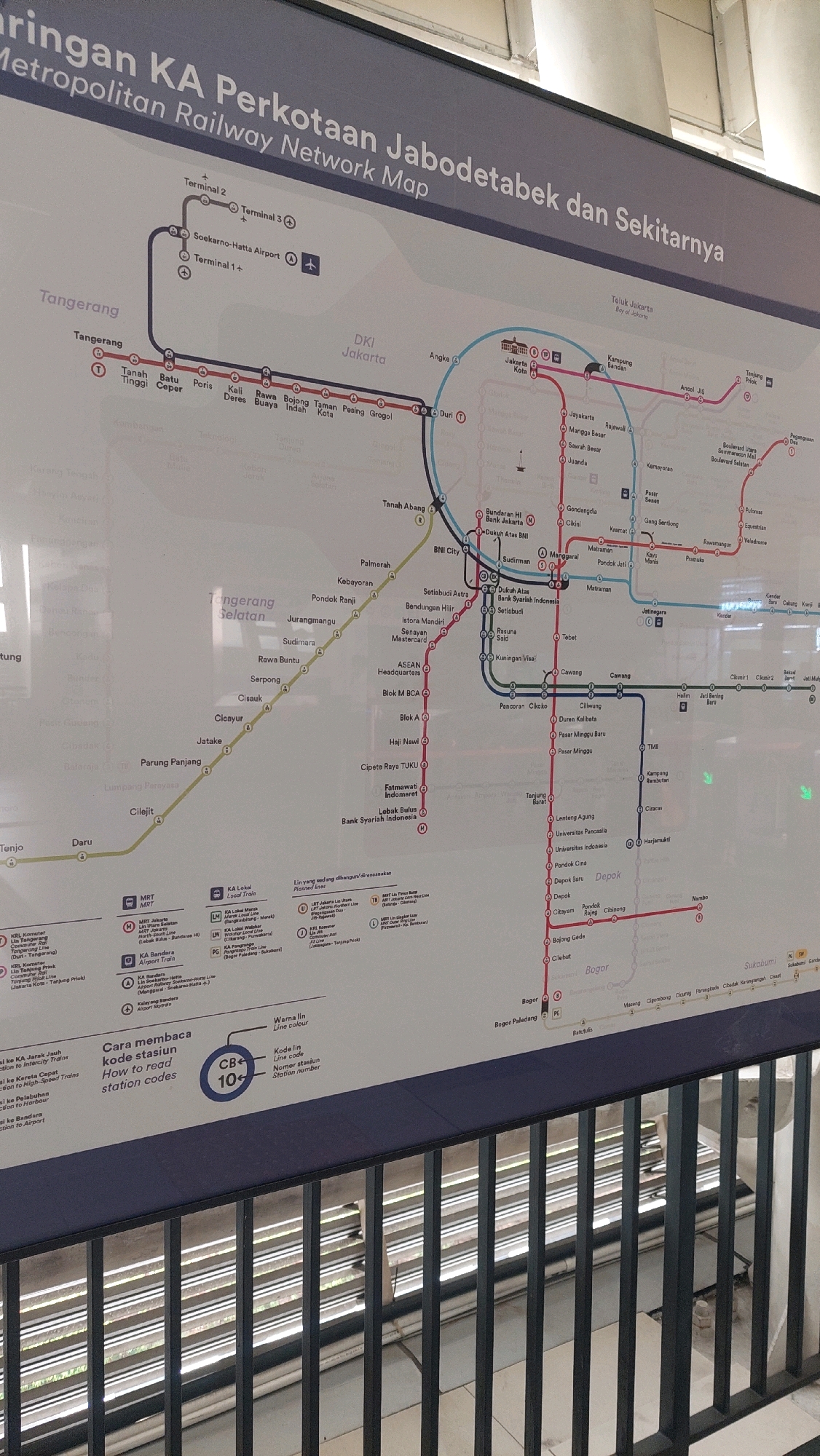 The image displays a detailed public transportation map of the Jabodetabek Metropolitan Railway Network in Jakarta, Indonesia. The map is presented on a large, vertical display screen or board, likely in a public transit station. The dominant colors are shades of blue, grey, and white for the background and text, with various colored lines representing different train routes: red, blue, yellow, and purple. Key locations and stations are labeled in both Indonesian and English, with some station names appearing in both scripts.
The map shows an intricate network of railway lines connecting various parts of the Jabodetabek region, including Jakarta, Tangerang, and Bogor. Several major transit hubs and airport connections are highlighted. There are also legends explaining the meaning of different line colors, station codes, and symbols. Specifically, there is a section titled "Cara membaca kode stasiun" (How to read station codes) which clarifies the meaning of line color and station number.
The map is mounted on a dark gray wall, with the bottom portion of the image showing the top of a metal railing, suggesting the photograph was taken from a vantage point above or in front of the railing, possibly on a walkway or platform overlooking the display. The lighting appears to be overhead and somewhat diffused, typical of an indoor public space. No people are visible in the immediate vicinity of the map, but the environment suggests a busy public transportation hub. The title of the map is "Jaringan KA Perkotaan Jabodetabek dan Sekitarnya | Metropolitan Railway Network Map". The presence of Jakarta and its surrounding areas confirms the geographical context.