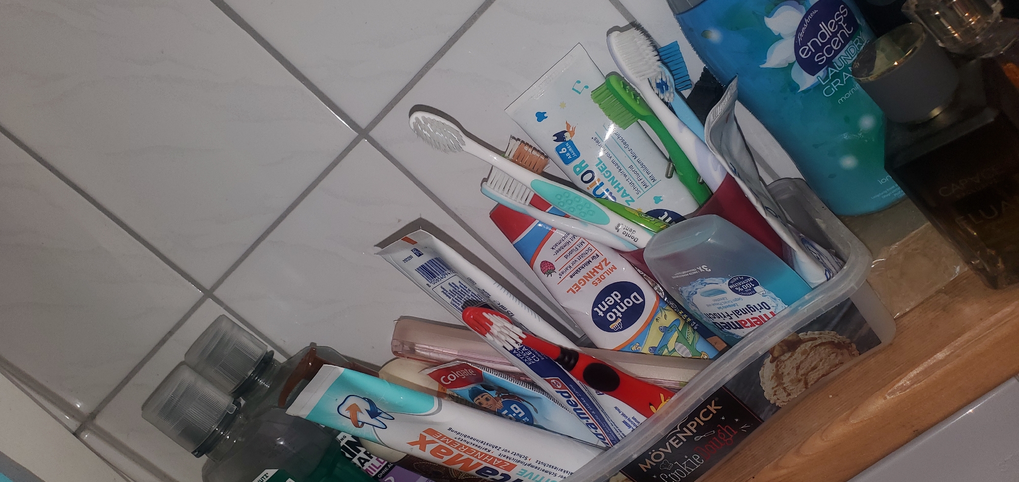 The image shows a close-up, slightly angled view of a collection of oral hygiene products and other items in a clear plastic container, resting on a wooden surface. The container is filled with various tubes of toothpaste, toothbrushes with different colored bristles, and possibly mouthwash bottles. Some visible brands and labels include "LaMax Zahncreme," "Colgate," "Donto Dent," "Junior Zahngel," "Mövenpick Cookie Dough," and "Endless Scent Laundry Granulate." The background consists of light-colored tiled walls. The lighting is somewhat dim, casting shadows and highlighting the textures of the objects. There are no people present in the image. The scene appears to be a bathroom or a similar personal care area. The image does not contain any identifiable landmarks or contextual clues that would suggest a specific location within Sasbach, Germany.