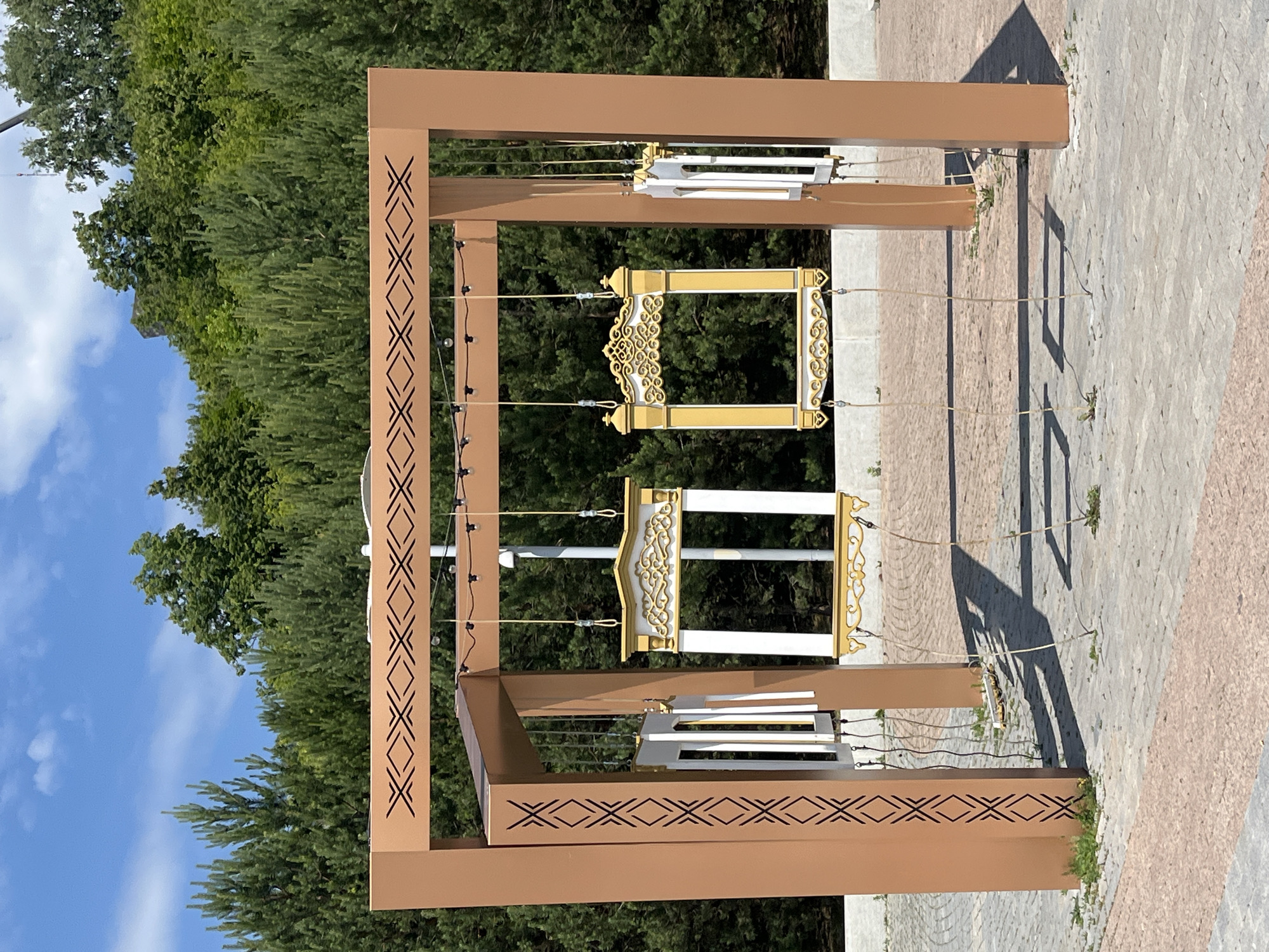 This is a vertical, eye-level shot of an outdoor art installation in Ufa, Russia, on a sunny day. The installation consists of a large, brown, rectangular frame supported by four posts. Decorative geometric patterns are carved into the vertical posts and the horizontal beam. Hanging from the horizontal beam by ropes are four decorative picture frames. Two of the frames are ornate and gold with white interiors, while the other two are plain white. They are suspended at varying heights, creating a sense of movement. The ground is paved with grey and pink interlocking bricks, with patches of green grass growing through. In the background, a dense forest of green trees rises up a gentle slope, under a bright blue sky with a few white clouds. There are no people visible in the frame.