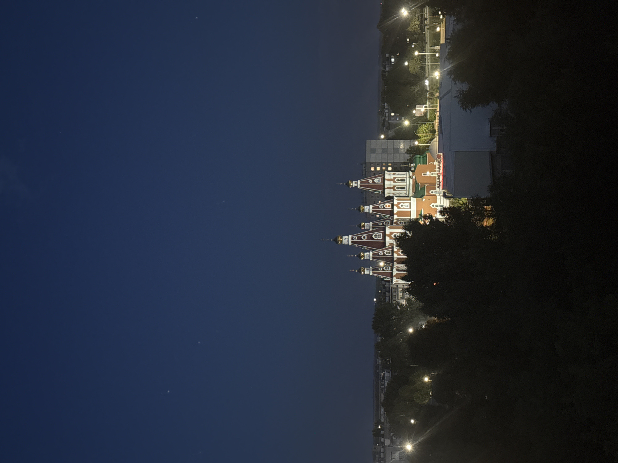 The image depicts an exterior view of a building with distinctive red and white onion domes, illuminated at night. The building appears to be a place of worship, possibly a church, given its architectural style. It is situated in a developed area with other buildings and streetlights visible in the background. Trees frame the right side of the image, creating a contrast between nature and the urban landscape. The sky is dark blue, indicating that it is nighttime, and there are a few faint celestial bodies visible. The lighting suggests a clear evening. The overall atmosphere is peaceful and serene, with the illuminated building serving as a focal point against the dark backdrop. There is some visible text on a building in the background, but it is not clearly legible.