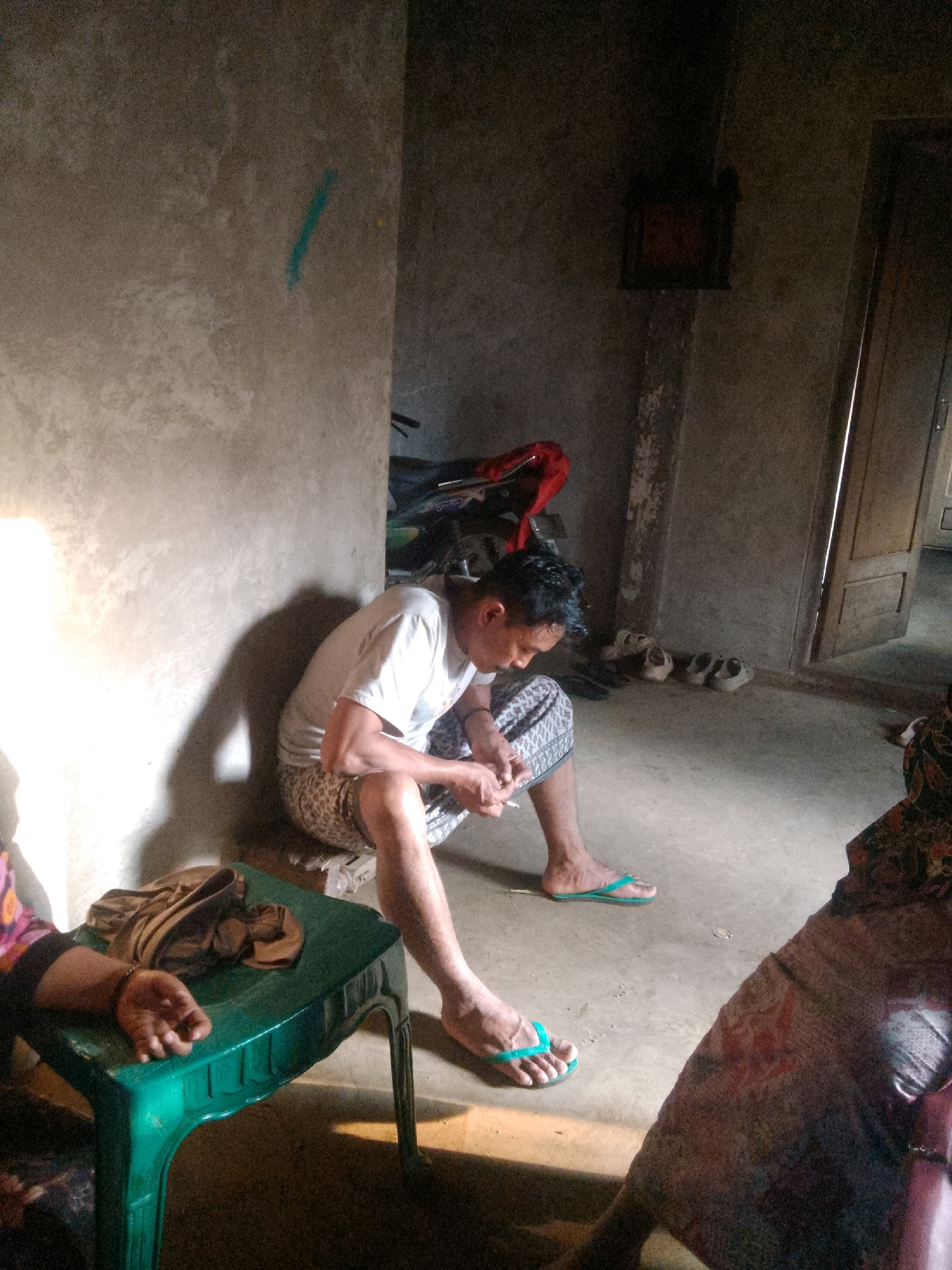 The image depicts an indoor scene, possibly in Krucil, Indonesia, where a man is seated on the floor, leaning against a wall. He is wearing a white t-shirt and patterned pants. His body is positioned in a way that suggests he is looking down at something in his hands. In front of him is a small, green plastic table with some brown fabric draped over it. To the left of the table, a person's arm and hand are visible, resting on the edge of the table. The floor is concrete and appears worn. In the background, a doorway is visible, and various objects are scattered around, including shoes and what appears to be a motorcycle. The lighting suggests it is daytime, with a strong light source coming from the left, casting a shadow of the man on the wall. The overall environment appears to be a modest, lived-in space.