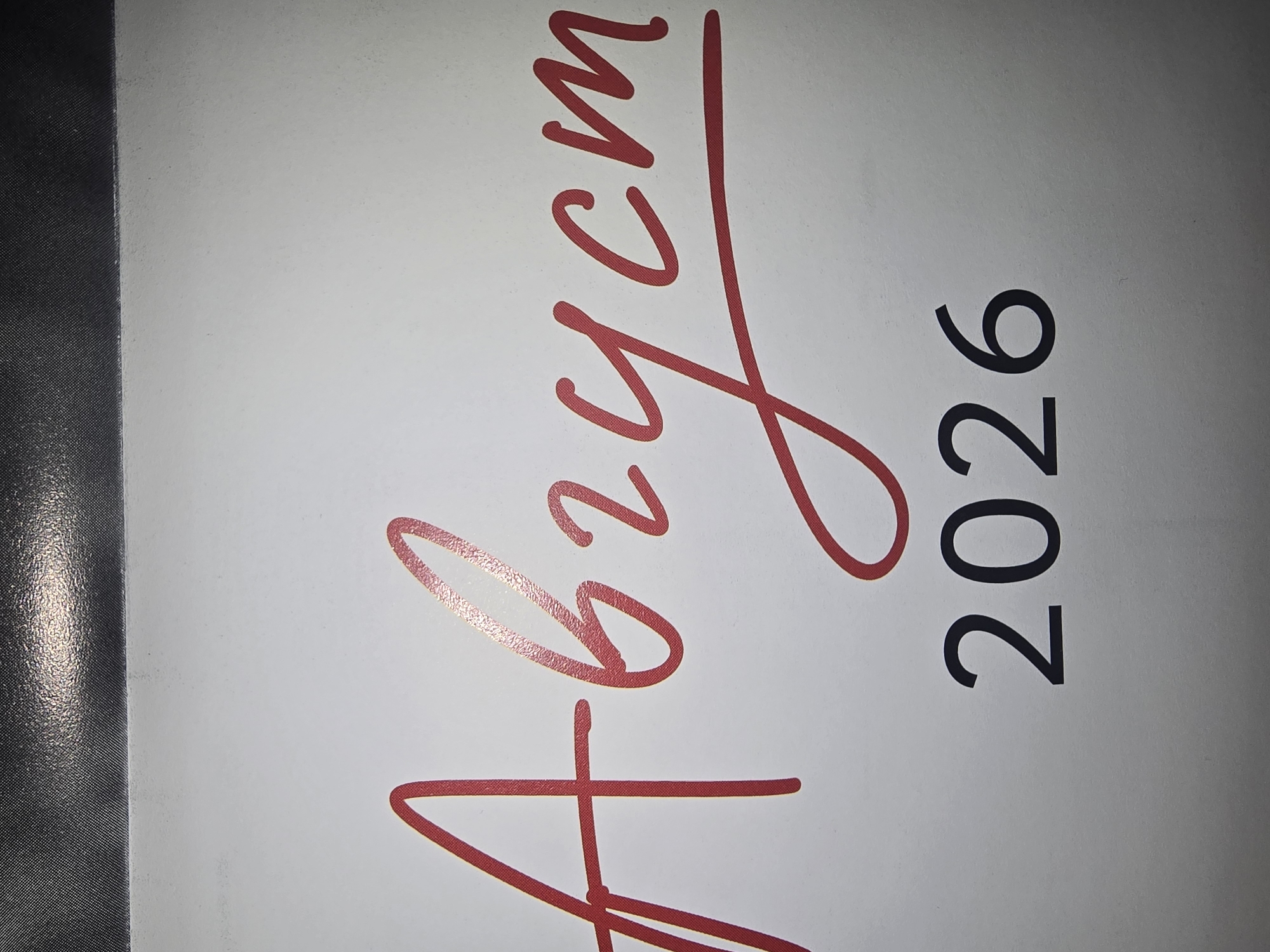 The image shows the word "Август" (August) written in a stylized red font, followed by the year "2026" in black sans-serif numerals. The text is centered on a white background. The top edge of the image displays a dark, textured band, possibly from the top of a calendar or a similar item. There are no people, specific locations in Ulan-Ude, or any actions depicted. The scene is a close-up of text on a plain surface.