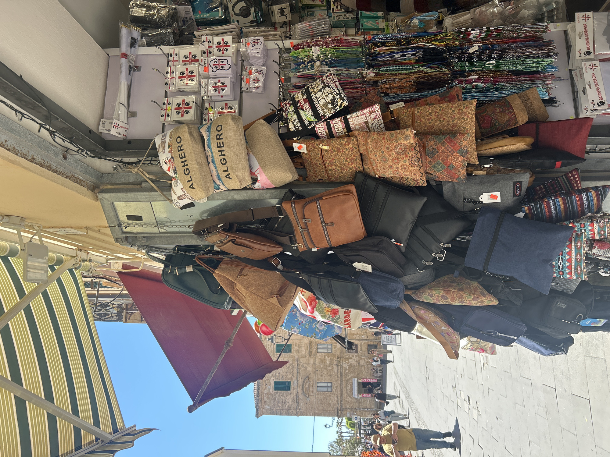 The image captures a bustling scene in Alghero, Italy, specifically at a market stall overflowing with merchandise. The stall's offerings include an array of bags, pouches, and accessories. Several items are labeled with "ALGHERO" or "Sardegna", indicating local souvenirs. There are strings of beads, small flags, and miscellaneous items.
Below the stall, pedestrians are visible, including a person wearing a yellow shirt. In the background, there is a building with multiple windows and an "Ice Cream" sign. An awning extends from the shop, providing shade. The scene suggests a vibrant, tourist-oriented atmosphere on a sunny day.