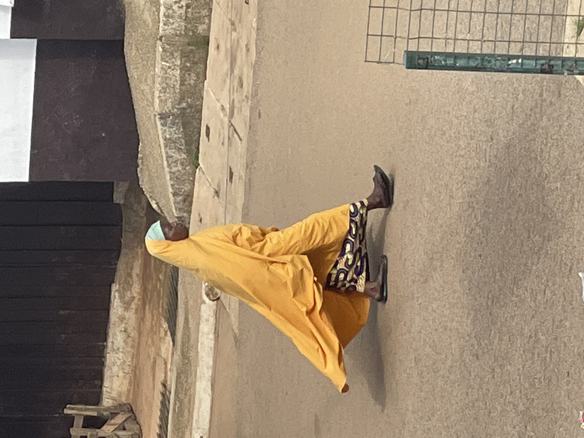 A person wearing a yellow robe and a light blue headscarf walks on a dirt path next to a stone building. The person's feet are visible, wearing dark sandals. The person is walking away from the camera, and their face is partially obscured by the robe. The setting appears to be outdoors, with a stone wall or building to the left and a sandy path to the right. A metal fence is visible in the background. The time of day and weather are unclear from the image. There is no visible text.