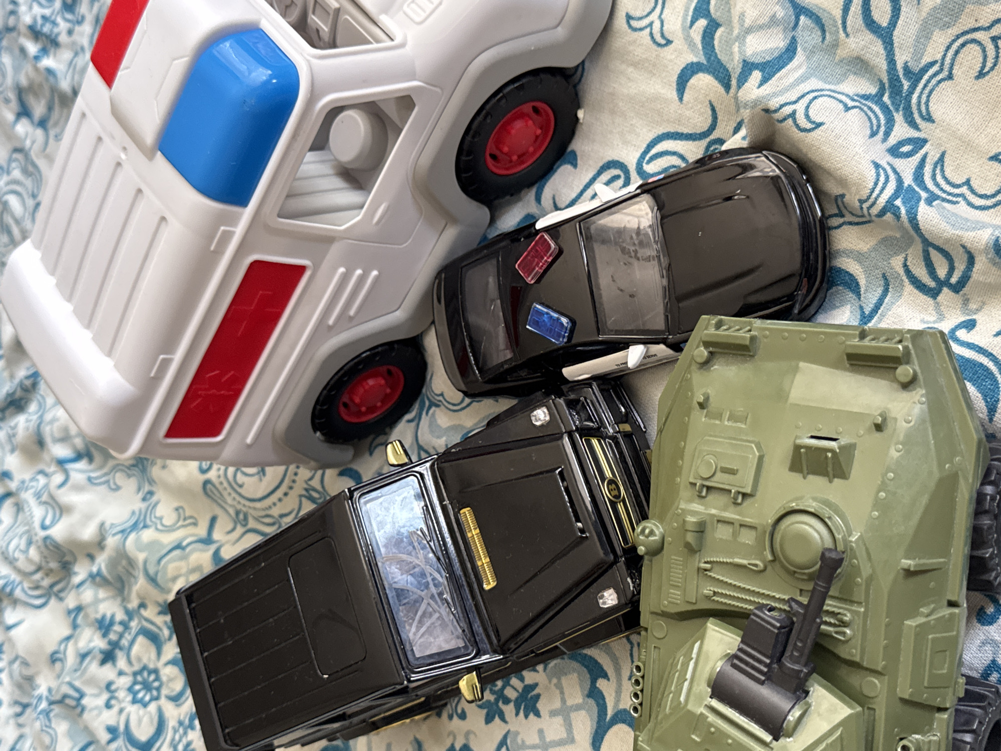 This is a close-up, eye-level, still shot of several toy vehicles on a patterned fabric surface. The vehicles include a green toy tank with visible treads and a turret, a black toy SUV with gold accents and tinted windows, a black toy police car with red and blue lights on the roof, and a white toy ambulance with red and blue lights. The fabric is white with a blue floral pattern. The scene appears to be an indoor setting, possibly a bedroom or playroom. There is no indication of the specific location within Яромінскі сельскі Савет, Belarus.