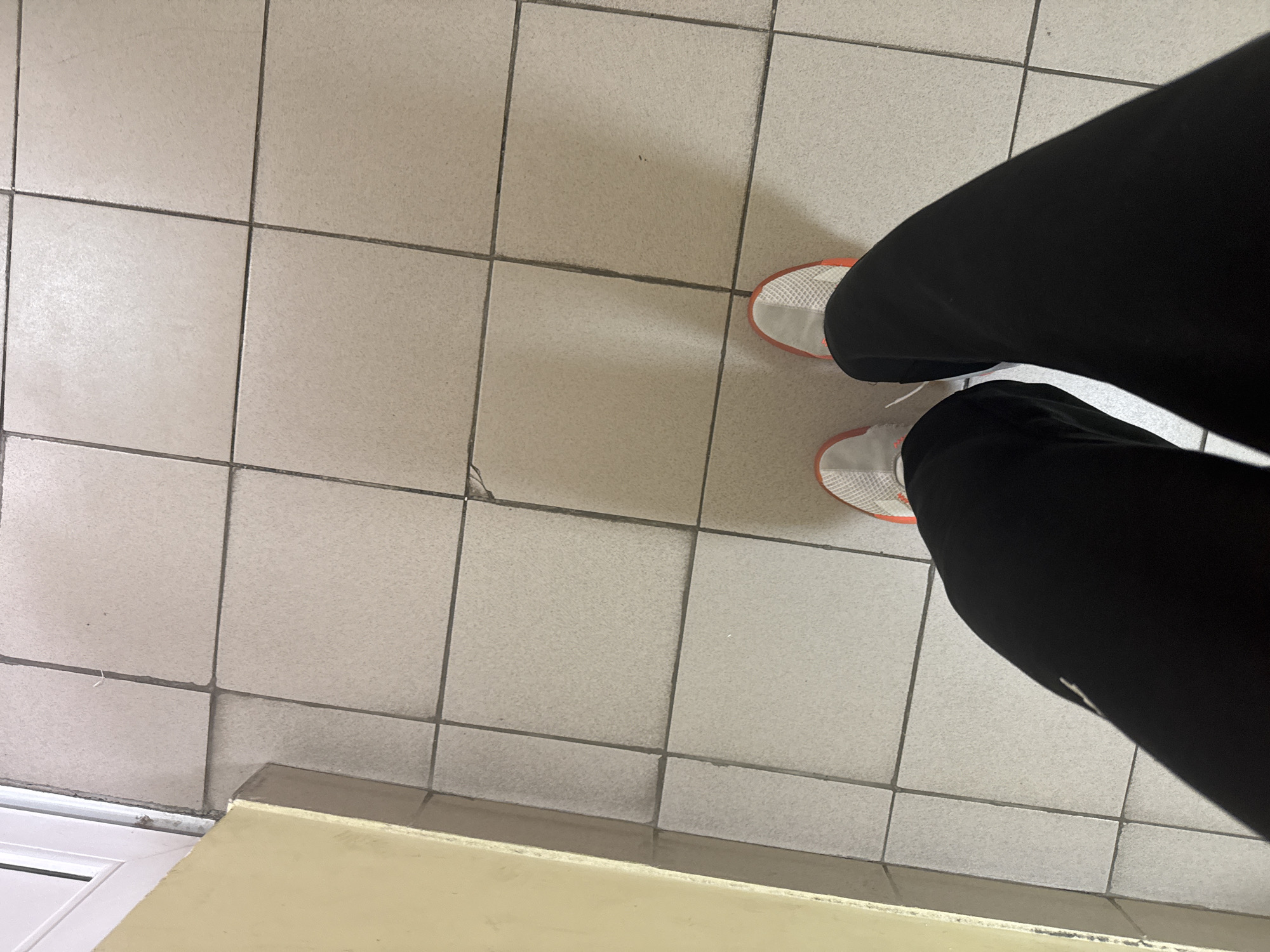 The image shows a top-down view of a person standing on a tiled floor. The person is wearing black sweatpants and white sneakers with an orange trim. The floor is made up of square tiles in a light gray color. There is a cream-colored baseboard or wall section visible in the lower left corner of the image. The location in the city of Kazan, Russia cannot be determined from the image.