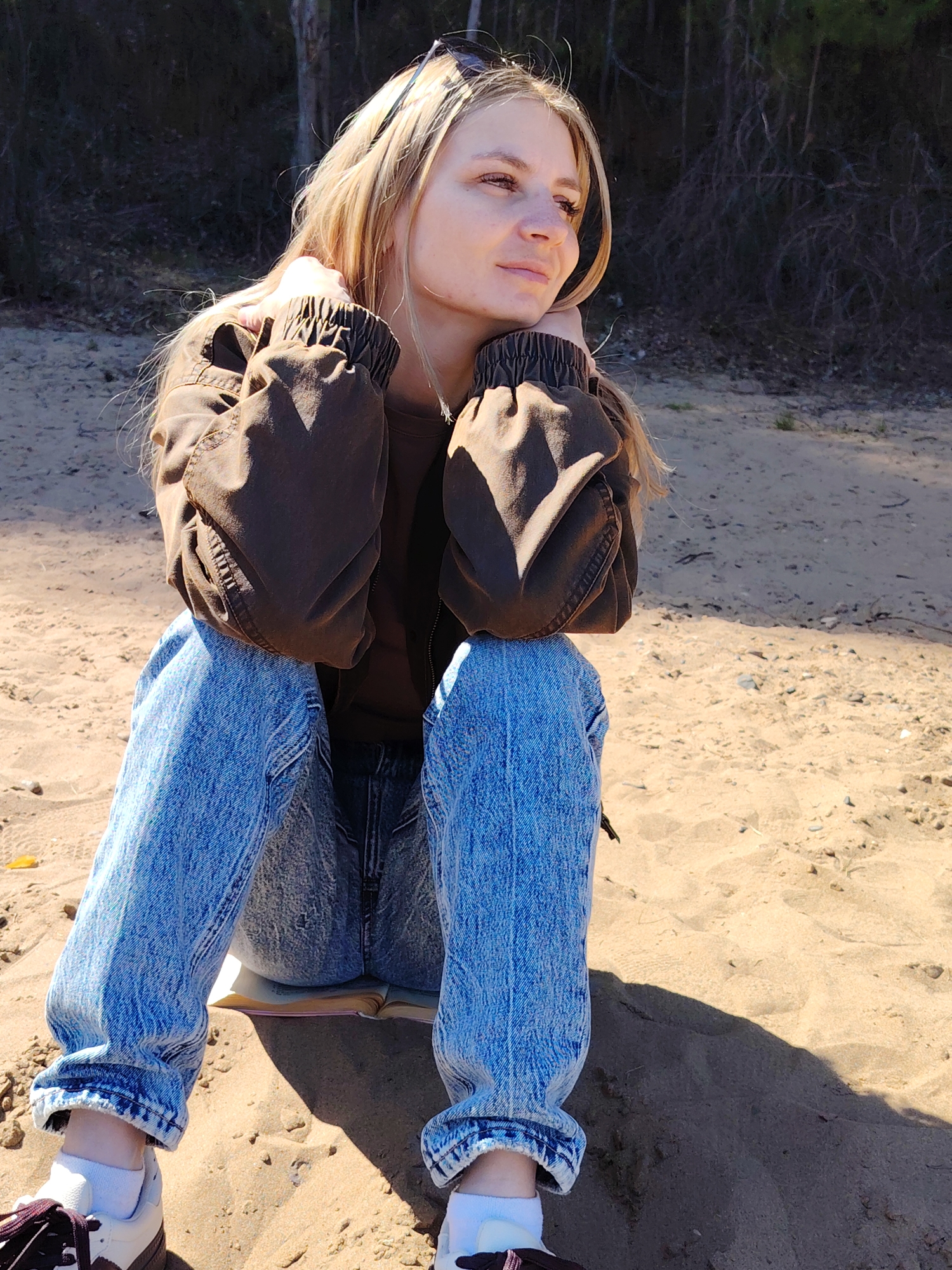 A young woman is seated on a sandy surface, presumably a beach, in an outdoor setting. She is wearing light blue jeans with the cuffs rolled up, brown and white sneakers with white socks, a brown long-sleeved shirt, and a brown bomber jacket. Sunglasses are perched on her head. She is sitting on an open book. Her legs are bent with her knees pointing upwards, and her arms are resting on her knees. She is looking off to the side, seemingly lost in thought. The background consists of trees and vegetation. The location is Chaikovsky, Russia.