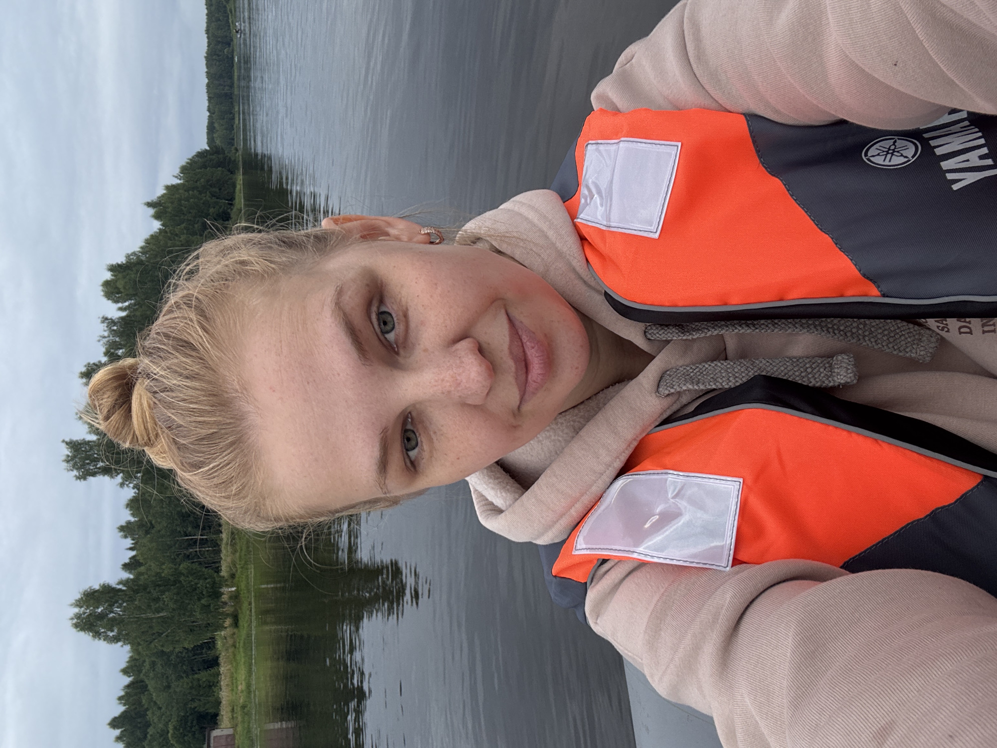 A young Caucasian woman with blonde hair pulled into a bun is taking a selfie on a body of water, possibly a river or lake. She is wearing an orange and grey life vest over a tan hoodie. The background shows a treeline with a cloudy sky overhead. A small building is visible on the left side of the frame. The scene appears to be in Anteplevo (Okhotinskoe S/P), Russia, given the context.