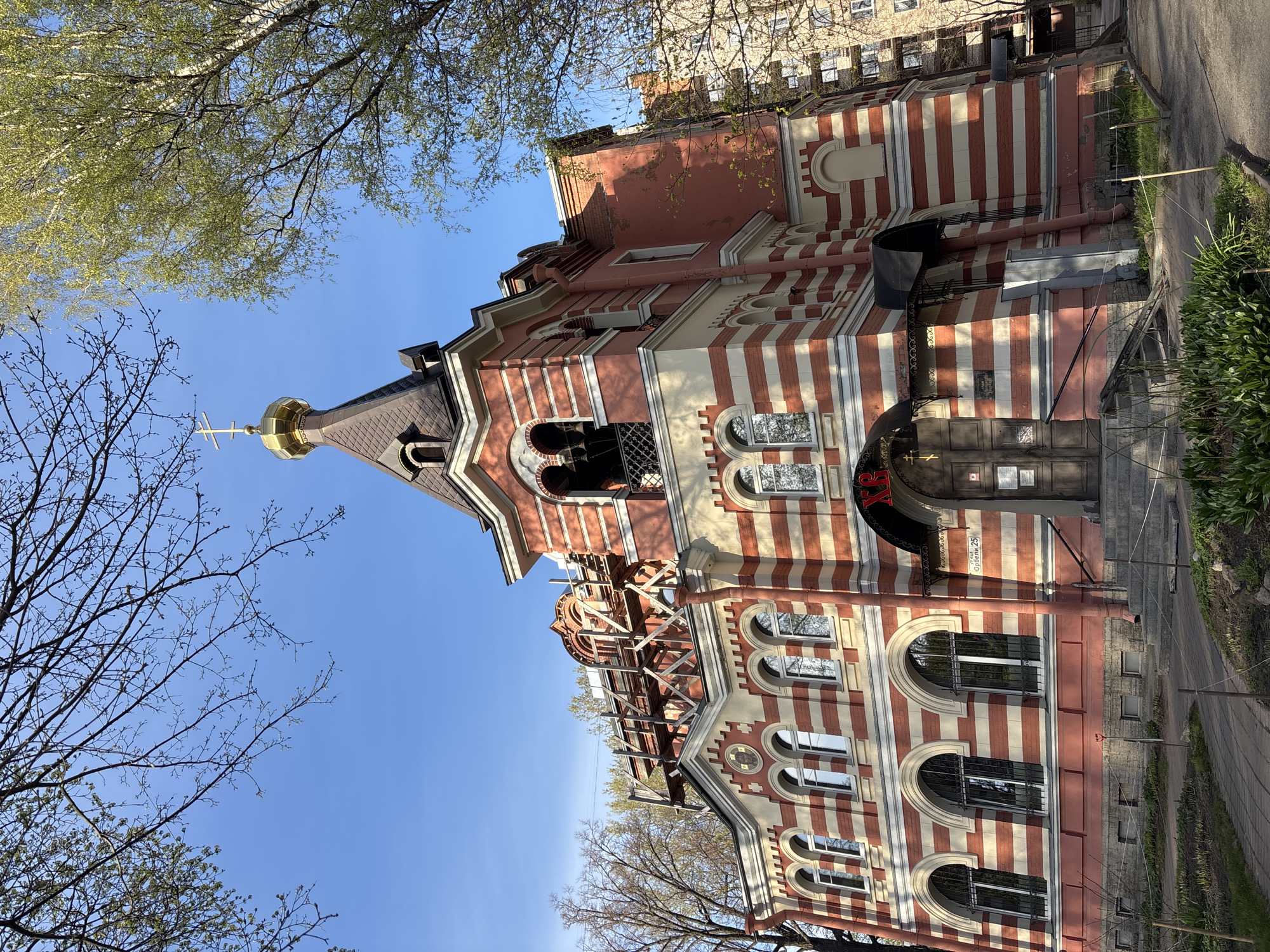 The image shows the Church of the Vladimir Icon of the Mother of God on Vladimirskaya Square in Saint Petersburg, Russia. The building has a distinct red and white striped facade with arched windows and a golden dome topped with a cross. Construction scaffolding is visible on the roof, indicating ongoing maintenance or renovation. The sky is blue, with tree branches framing the scene.