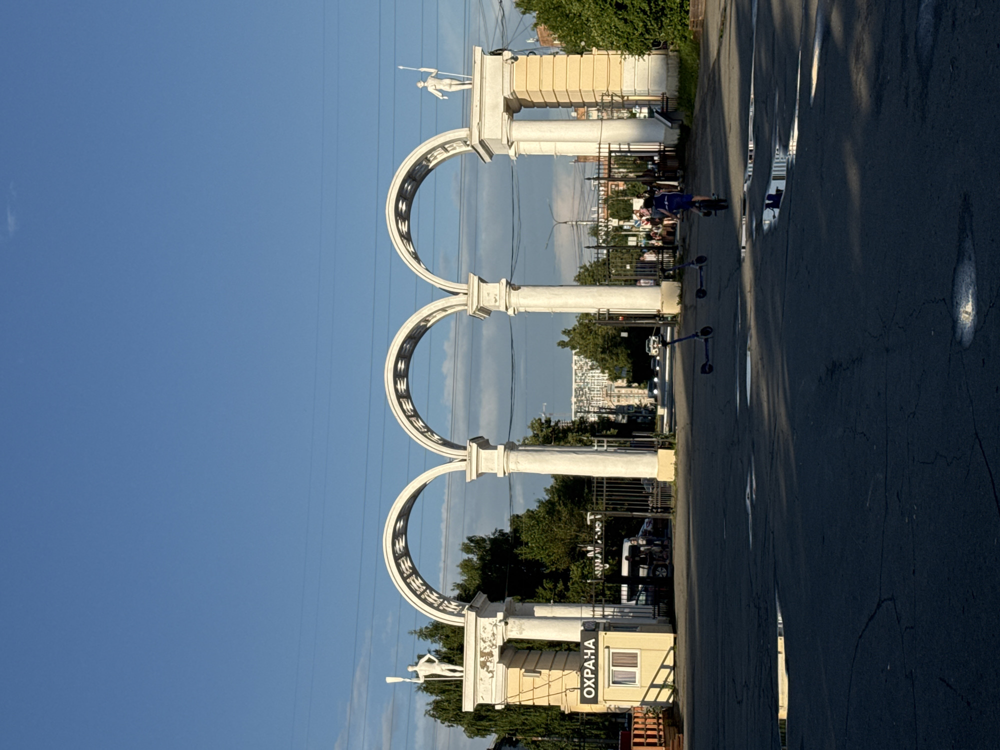 The image depicts a large, white, three-arched gateway in Izhevsk, Russia, likely an entrance to a park or administrative area. The gateway features classical architectural elements, with tall columns and statues of male figures holding staffs atop the outer pillars. A small yellow security booth with the word "ОХРАНА" (Security) written on it is situated to the left of the entrance.
The scene is set on a paved road with several puddles reflecting the sky and surrounding environment. In the foreground, three electric scooters are parked or in use. One person, wearing a blue outfit, is riding a scooter away from the camera. In the background, through the arches and beyond a metal fence, a street scene is visible with cars and pedestrians. The environment is illuminated by natural daylight, under a clear blue sky with some faint clouds. Trees and buildings are visible on either side of the gateway, suggesting an urban setting.
