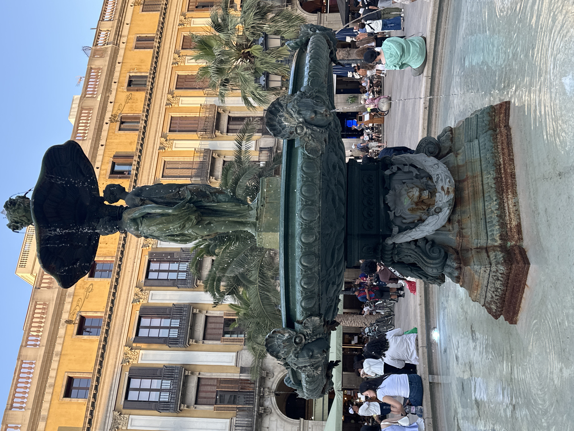 This is a full shot of a large, ornate, bronze fountain in a public square in Barcelona, Spain. The fountain has multiple tiers, with the top featuring a large basin and sculptural figures. Water flows from the top basin down into a lower, wider basin, which is partially filled with water. The fountain is situated in a paved square with several people gathered around it.
In the background, there is a multi-story yellow building with arched windows and balconies, indicative of European architecture. Palm trees are visible behind the fountain and in front of the building. People are seen sitting on the edge of the fountain, walking in the square, and sitting at outdoor cafes. Bicycles are also visible, suggesting a mode of transport or leisure activity in the area. The overall lighting suggests it is either early morning or late afternoon, with a clear blue sky.
The fountain itself is detailed with sculpted faces and decorative elements, showing signs of age and patina. The water in the fountain is clear, with ripples on the surface. The people in the scene are casually dressed and appear to be enjoying the public space. The scene is one of a lively urban environment with a prominent artistic centerpiece.