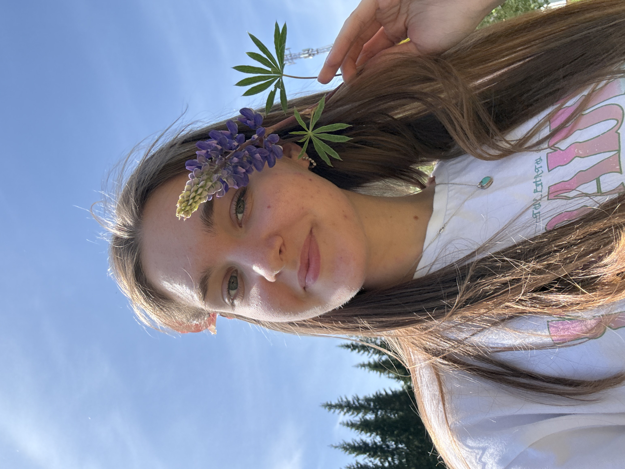 A young woman is shown in a close-up selfie. She has long brown hair and a lupine flower arrangement tucked behind her ear. The flower arrangement consists of purple flowers and green leaves. She is wearing a white t-shirt with a pink design, and a silver necklace with a turquoise pendant. The background is a clear blue sky and some green trees. The location is inferred to be Sibir', Russia based on the presence of lupine flowers which are common in the region.