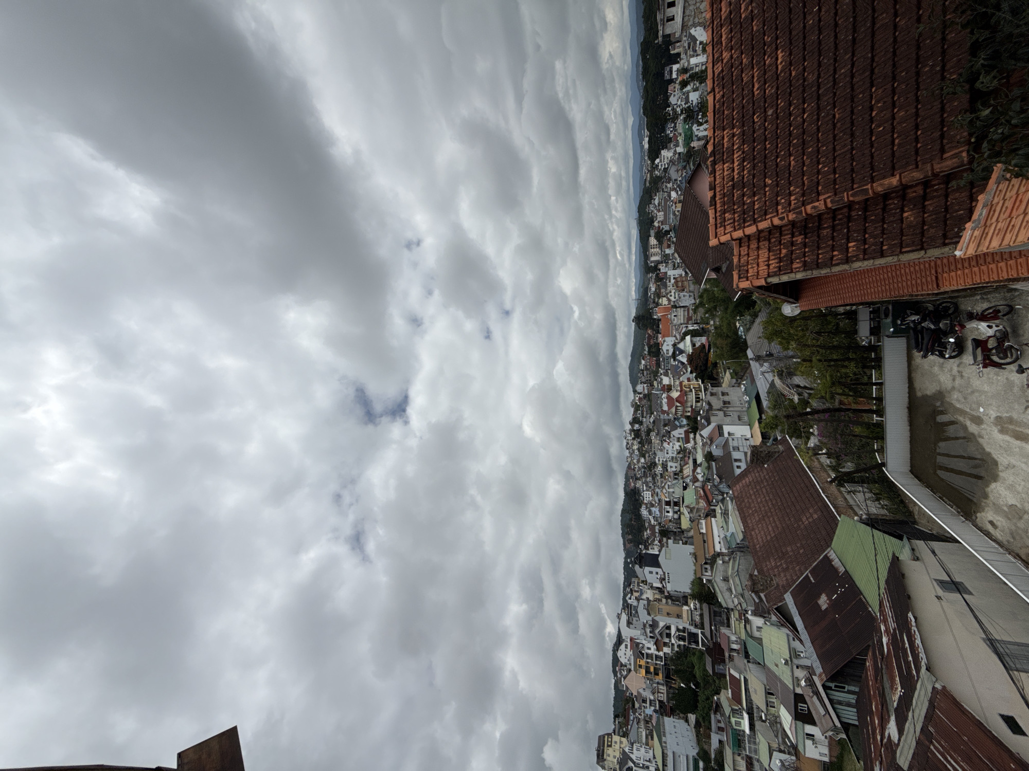 The image captures a panoramic view of Da Lat, Vietnam, under a cloudy sky. The foreground features the tiled rooftops of several buildings, some with rusted metal roofs. To the left, a portion of a darker building structure is visible. In the middle ground, a dense collection of houses and buildings sprawls across rolling hills, characteristic of Da Lat's hilly terrain. The architecture varies, with many houses featuring colorful facades and traditional tiled roofs. In the background, more distant hills and a hazy horizon are visible. Several motorcycles are parked in an open area in the lower right quadrant of the image, suggesting this is a residential or commercial area. The overall impression is of a bustling urban landscape nestled within a natural, hilly environment.