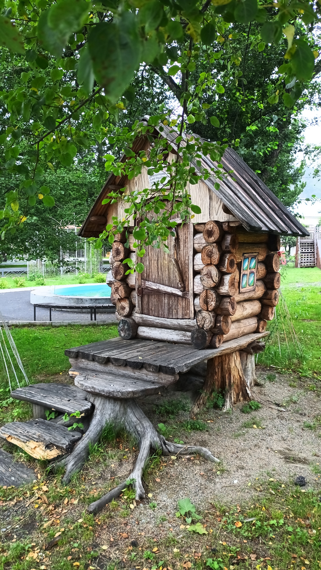This is a medium shot of a small, rustic wooden playhouse resembling a log cabin, set in a grassy outdoor area. The playhouse is built on tree stumps and features a wooden door and a small window with stained glass panes. A wooden platform extends from the front of the playhouse, with a set of rough-hewn wooden steps leading up to it. The structure is surrounded by green grass, fallen leaves, and tree roots. In the background, there's a glimpse of a blue swimming pool or wading pool, a fence, and trees. The overall atmosphere is whimsical and geared towards children's play. The lighting suggests it is daytime, and the presence of green leaves on the trees indicates it is likely spring or summer. A sign on the playhouse reads "Скворечник для тебя" which translates to "Birdhouse for you".