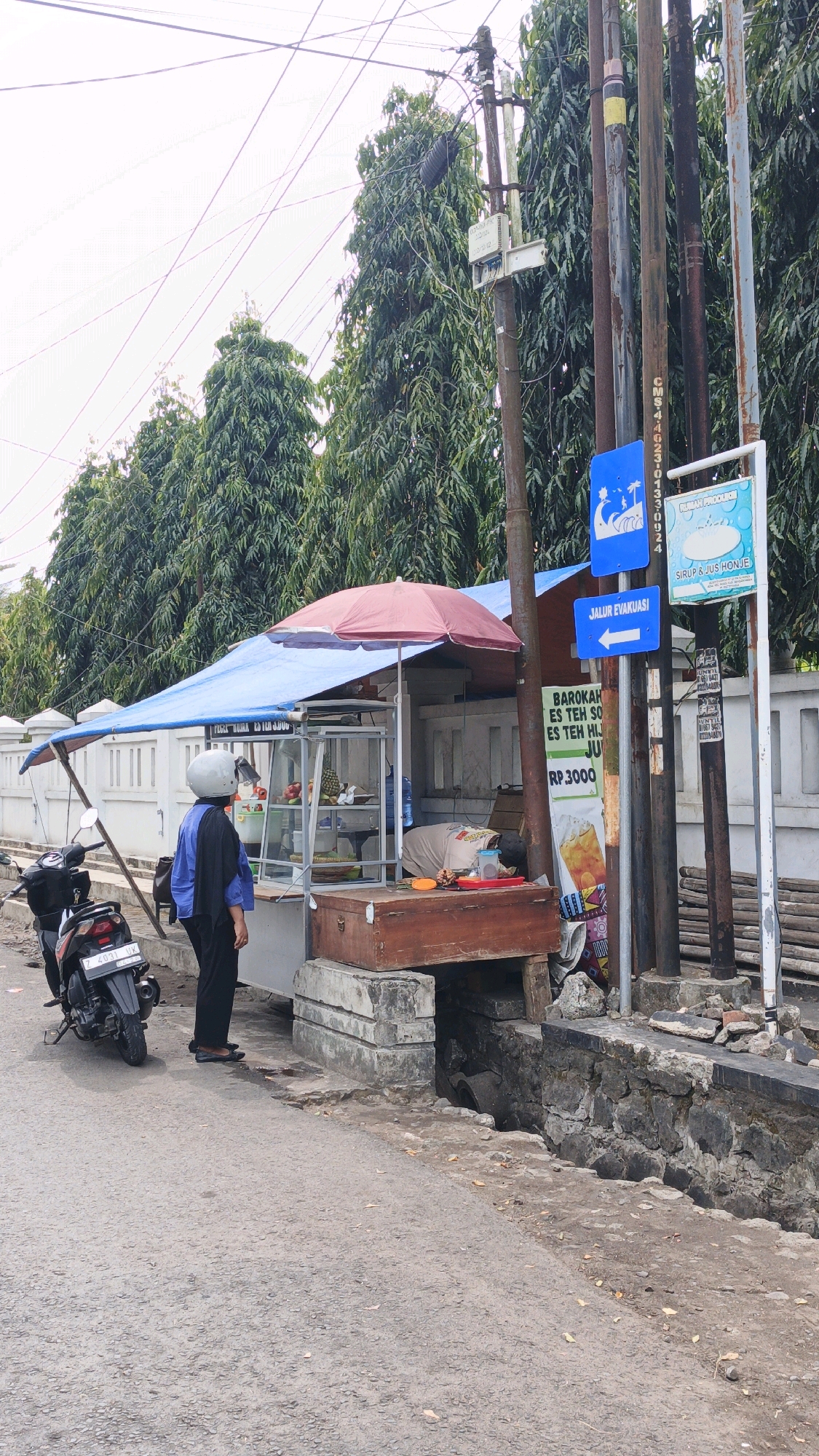 The image depicts a street scene in Desa Karangbenda, Indonesia. In the foreground, a paved road with some debris is visible. To the right, a trench with stone retaining walls runs alongside the road. A street food stall with a blue tarp roof and a red umbrella is set up on the side of the road. Display cases inside the stall contain fruits and bottles. A person wearing a blue shirt and black pants, with a white helmet on their head, stands looking at the stall. Another person is lying down on a wooden platform behind the stall, appearing to be asleep or resting. A motorcycle is parked to the left of the stall. Several utility poles with wires and various signs, including a blue sign with an evacuation route arrow, are present. In the background, a line of tall, leafy trees forms a dense green backdrop, and a white fence is visible behind the stall. The sky is overcast.