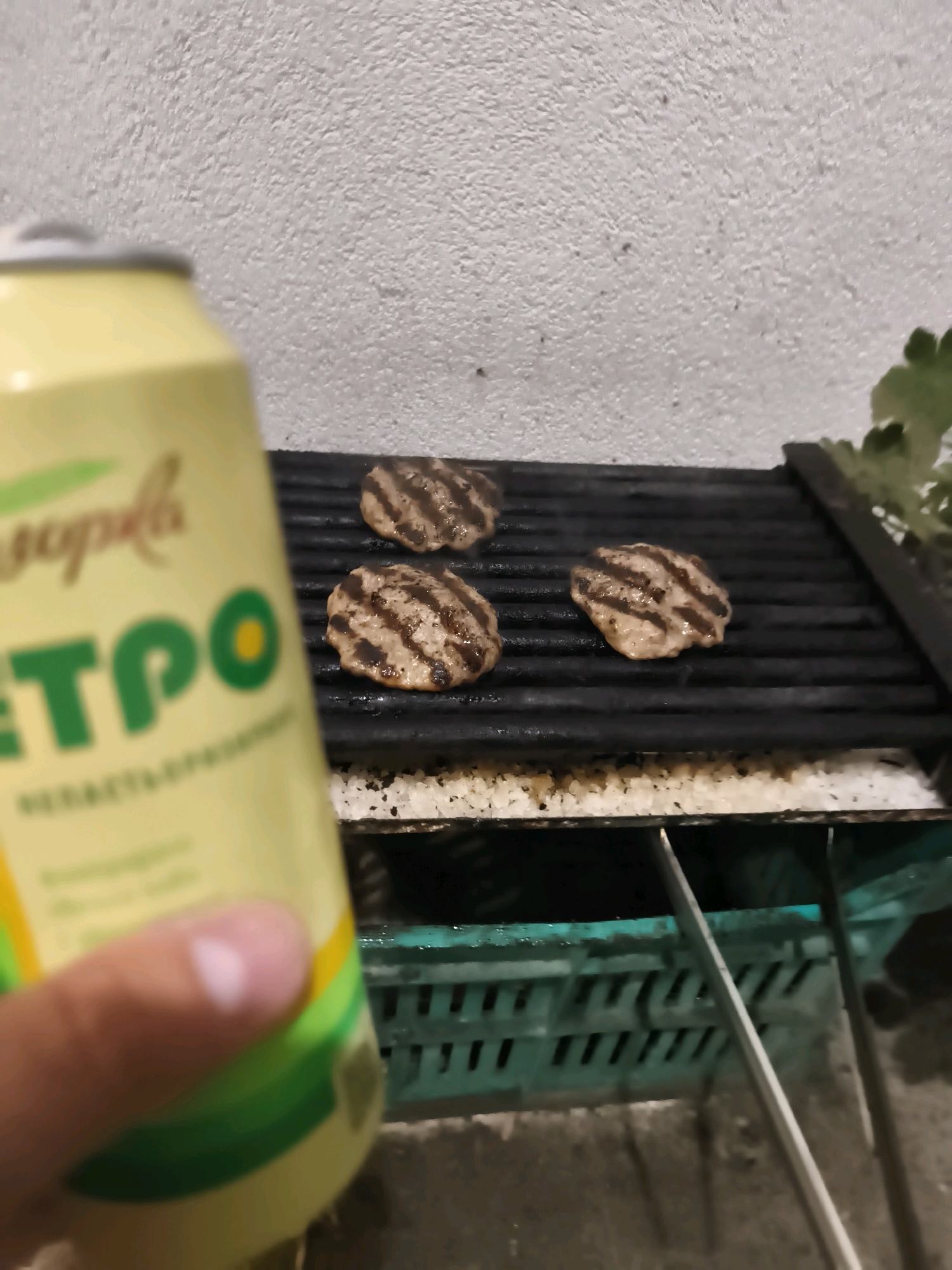 A can of soda is in the foreground, held by a person's hand, obscuring a grill with three burgers cooking on it. The grill is black with horizontal grates. Below the grill is a green plastic basket. In the background, a white textured wall is visible. The lighting suggests it is nighttime or indoors.