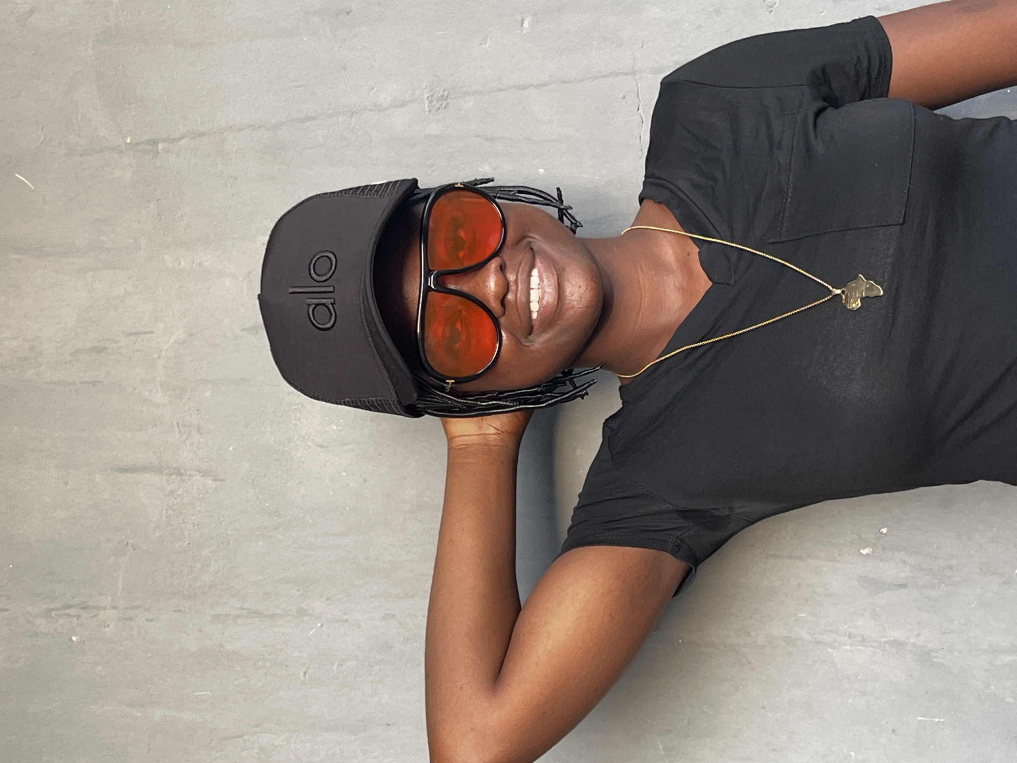 A woman is lying on a gray surface, likely concrete, with her head tilted to the left. She is wearing a black t-shirt, a gold necklace with a butterfly pendant, a black cap with "alo" printed on the front, and sunglasses with red lenses. Her right arm is bent and resting under her head. She is smiling. The image does not provide information to confirm the location in Badagry, Nigeria.
