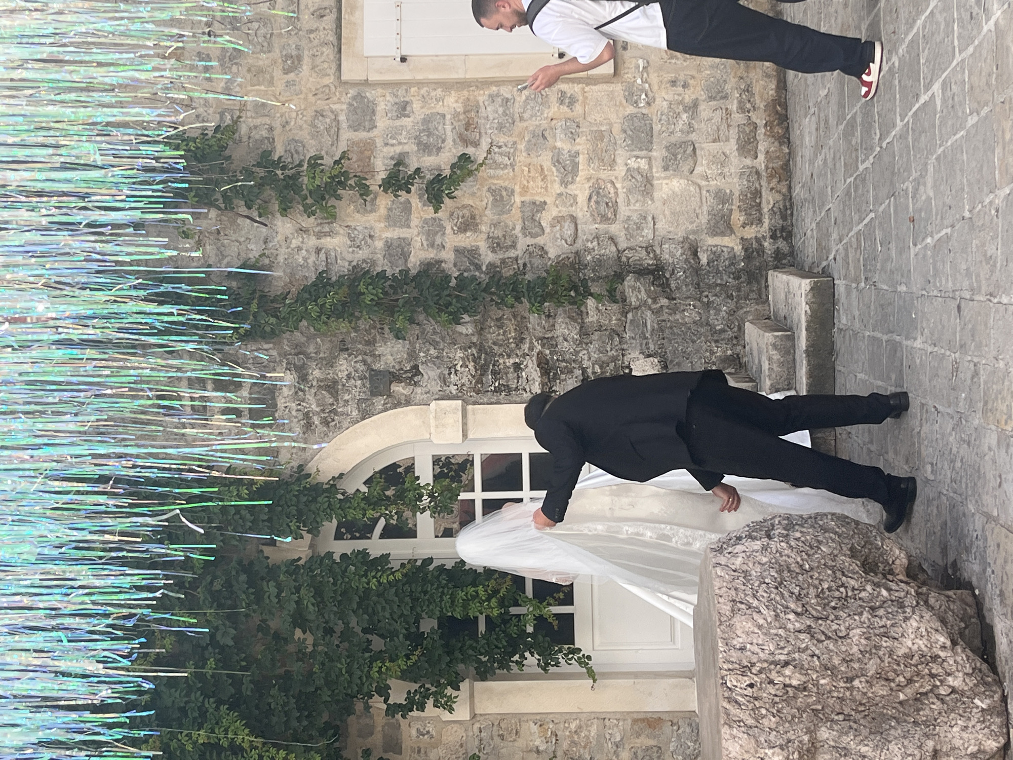 An outdoor scene in what appears to be a historic stone setting, possibly in Budva, Montenegro. On the left, a vibrant display of iridescent, shimmering streamers hangs vertically, suggesting a festive decoration. The background consists of an old stone wall, adorned with climbing green vines or ivy.
In the upper right portion of the image, a man stands on a paved stone path. He wears a white short-sleeved shirt, a dark vest or apron, dark trousers, and red and white sneakers. He is seen holding and looking down at a smartphone. Behind him, a window with white closed shutters is set into the stone wall.
In the lower right, closer to the viewer, another man in a black suit and black dress shoes is bending down. He appears to be assisting a woman, visible only by her elaborate white wedding dress and long, flowing white veil. His hand is near her dress, suggesting he is adjusting it. They are positioned in front of an arched window with white frames, also surrounded by green foliage. A large, rough-textured stone block sits prominently in the foreground on the right.
The lighting indicates daytime, with bright but not overly harsh sunlight. The overall atmosphere suggests preparations for a special event, likely a wedding, given the attire and decorations. No visible text is present in the image.