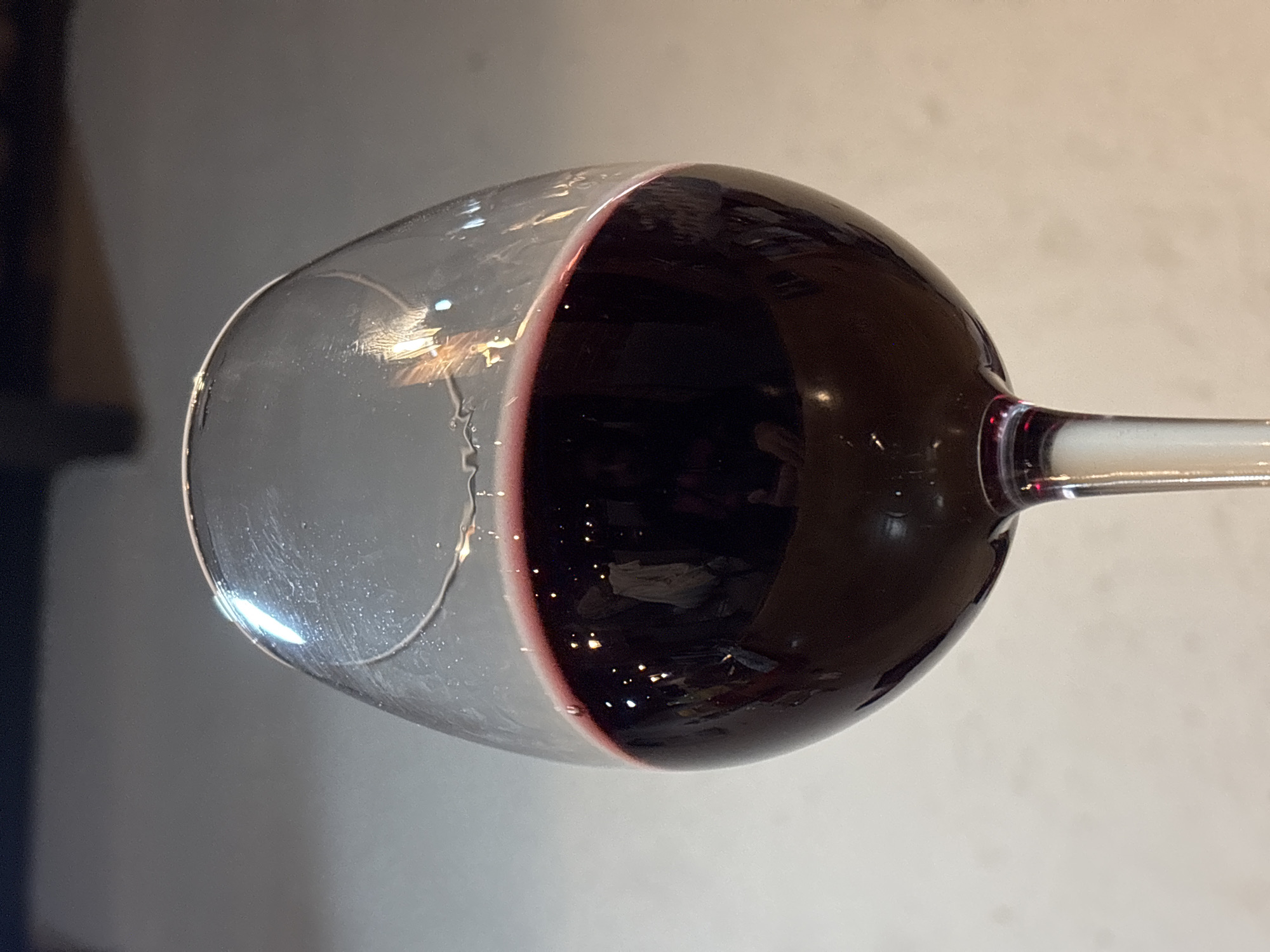 A close-up, tilted shot of a partially filled wine glass dominates the image. The left side of the glass is empty and clear, showcasing a slight interior condensation or residue. The right side is filled with a dark red liquid, presumably red wine, showing reflections of light. The glass has a thin, clear stem, extending to the right. The background is a blurred mix of light and dark tones, with a dark object on the left side, possibly furniture. The overall setting appears to be indoors. There is no evidence to determine the location as Puebla, Mexico. No people or interactions are directly visible in the frame.