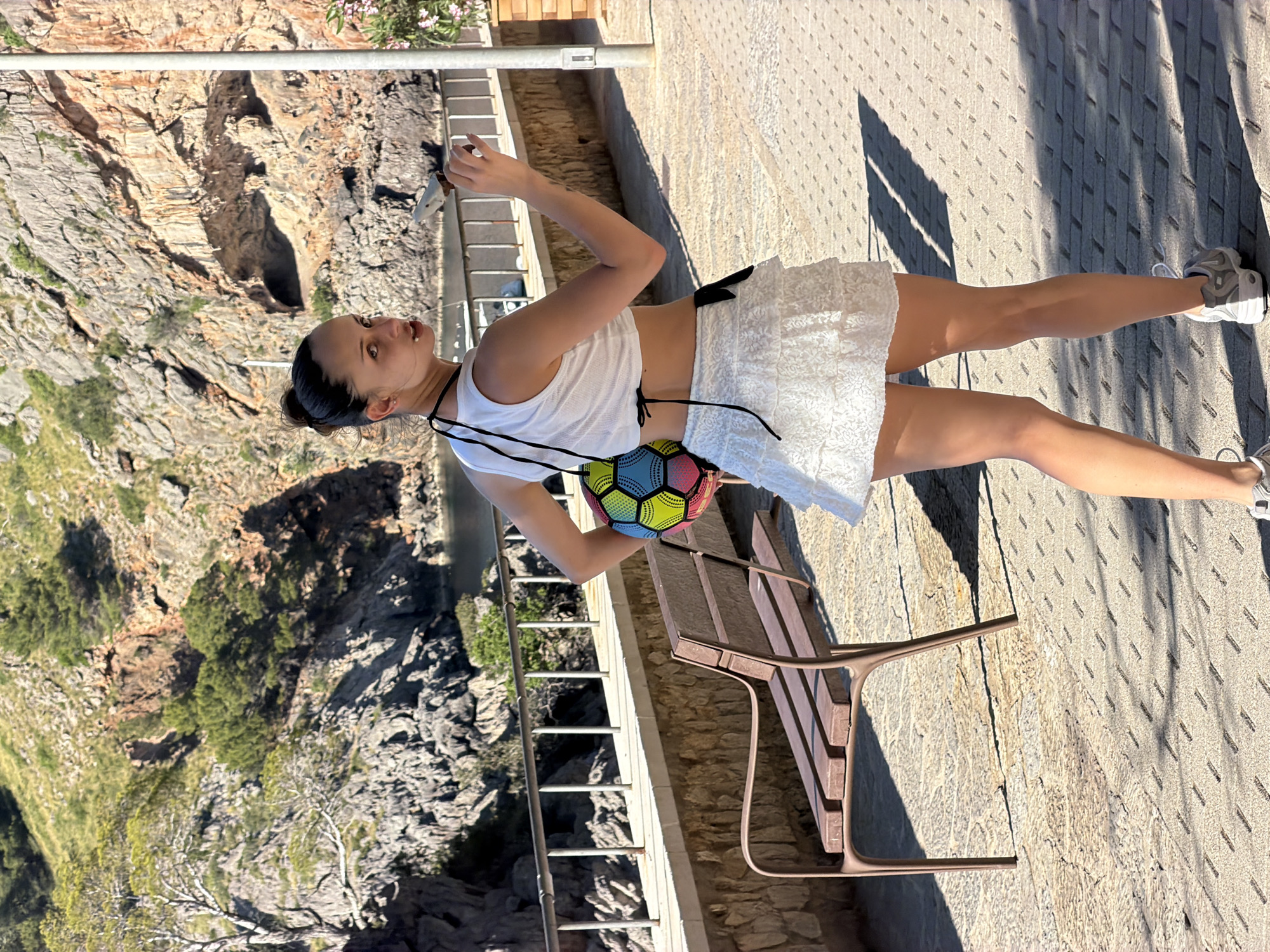 A woman wearing a white tank top and white skirt stands on a paved walkway next to a bench, looking towards a rocky cliff. She carries a colorful ball-shaped bag. The location is the city of Escorca, Spain.