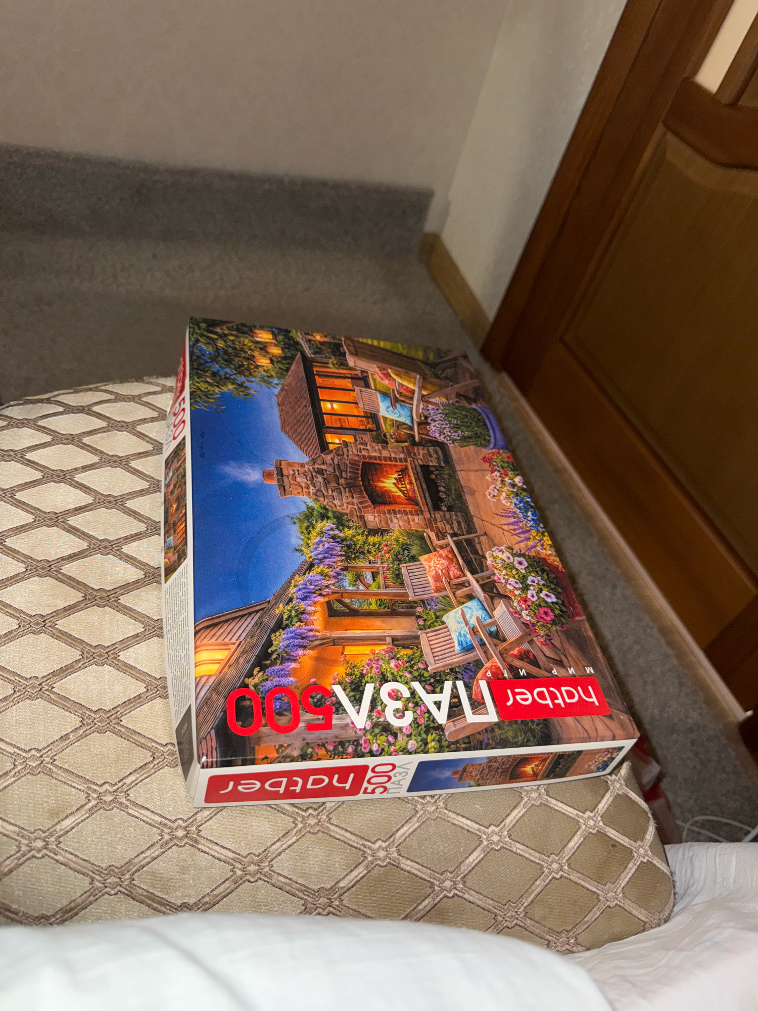 A jigsaw puzzle box, depicting a cozy evening scene with a fireplace and garden, is resting on a textured surface, possibly a cushioned seat or bed, in an indoor environment. The puzzle is identified as "HATBER 500 ПАЗЛ". The background includes a carpeted floor and a wooden door frame. The location is not specified as Zavolzhye, Russia, as there are no identifiable landmarks or clues to support this.