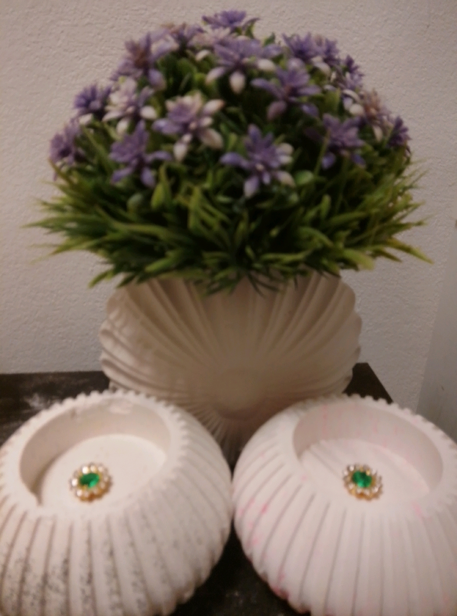 The image shows a close-up view of a decorative arrangement. In the center, a cream-colored, shell-shaped vase holds an artificial arrangement of green foliage and small purple and white flowers. In front of the vase, on a dark surface, are two white, round decorative bowls. Each bowl has a ridged exterior and a scalloped rim. In the center of each bowl, there is a small, ornate embellishment featuring a green stone surrounded by smaller clear or white stones, set within a gold-colored frame. The background is a textured, off-white wall. The image does not provide any information about the city of Wedel, Germany, or any specific location within it.