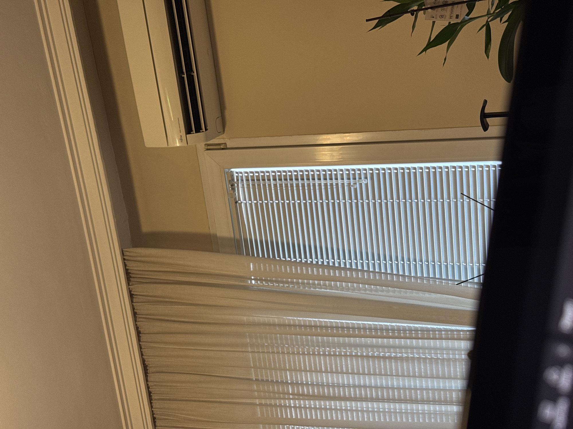 The image shows a window with white blinds, partially covered by a sheer white curtain. An air conditioning unit is mounted above the window on a light beige wall. A green plant is visible in the upper right corner. A dark object obstructs the view on the right side of the image. The location of the image is uncertain, but it's claimed to be in Sofia, Bulgaria.