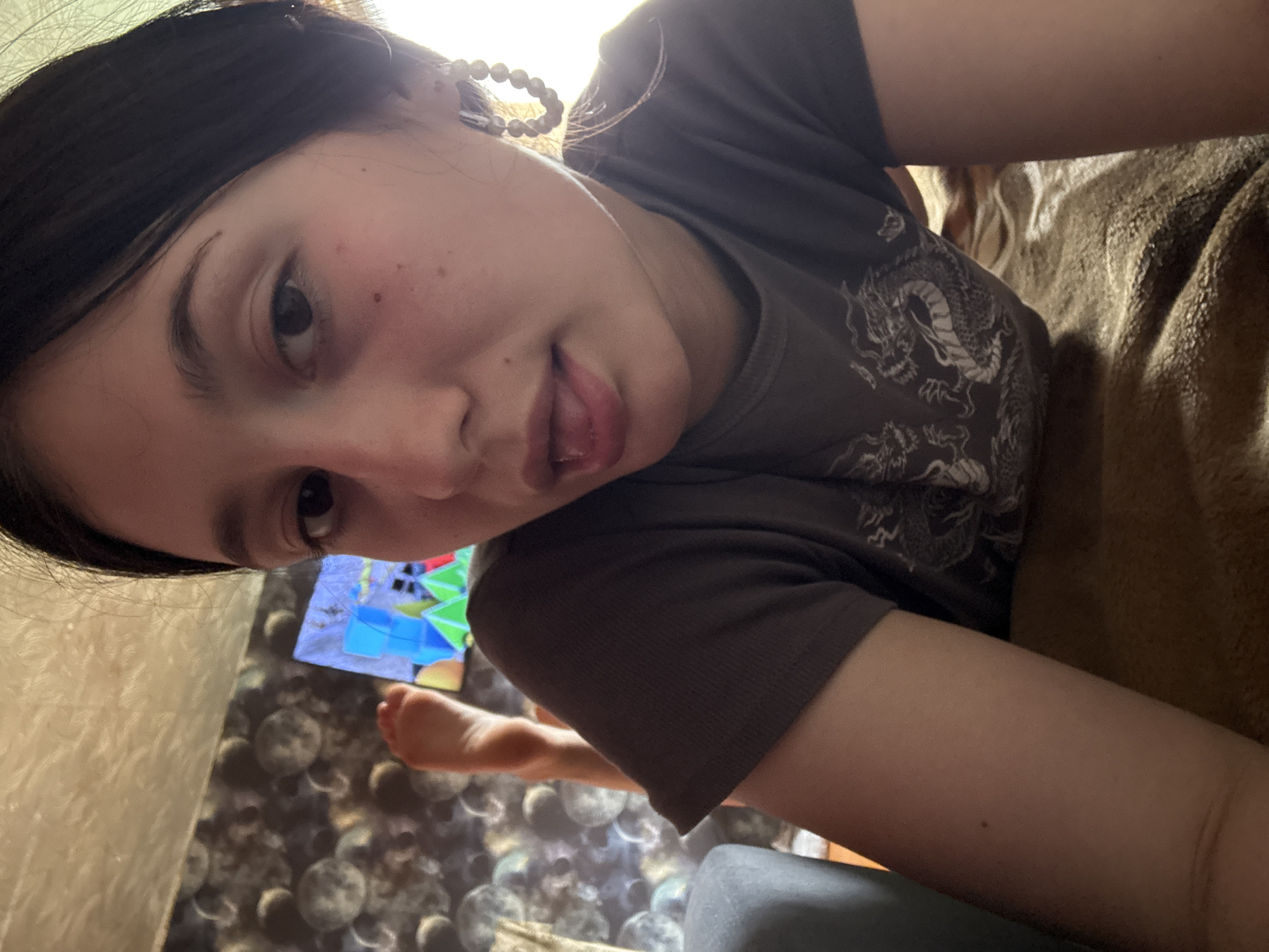 In a dimly lit room with textured wallpaper featuring a circular pattern, a young woman with dark hair and light skin is lying on a bed. She is wearing a dark t-shirt with a dragon graphic and large hoop earrings with pearl-like beads. Her right leg is propped up, with her bare foot visible. A television screen displaying colorful geometric shapes is positioned in the background. The overall setting suggests a casual, indoor environment, possibly a bedroom or living area in a home. The specific location within the city of Fokino, Russia, cannot be determined from the provided image.